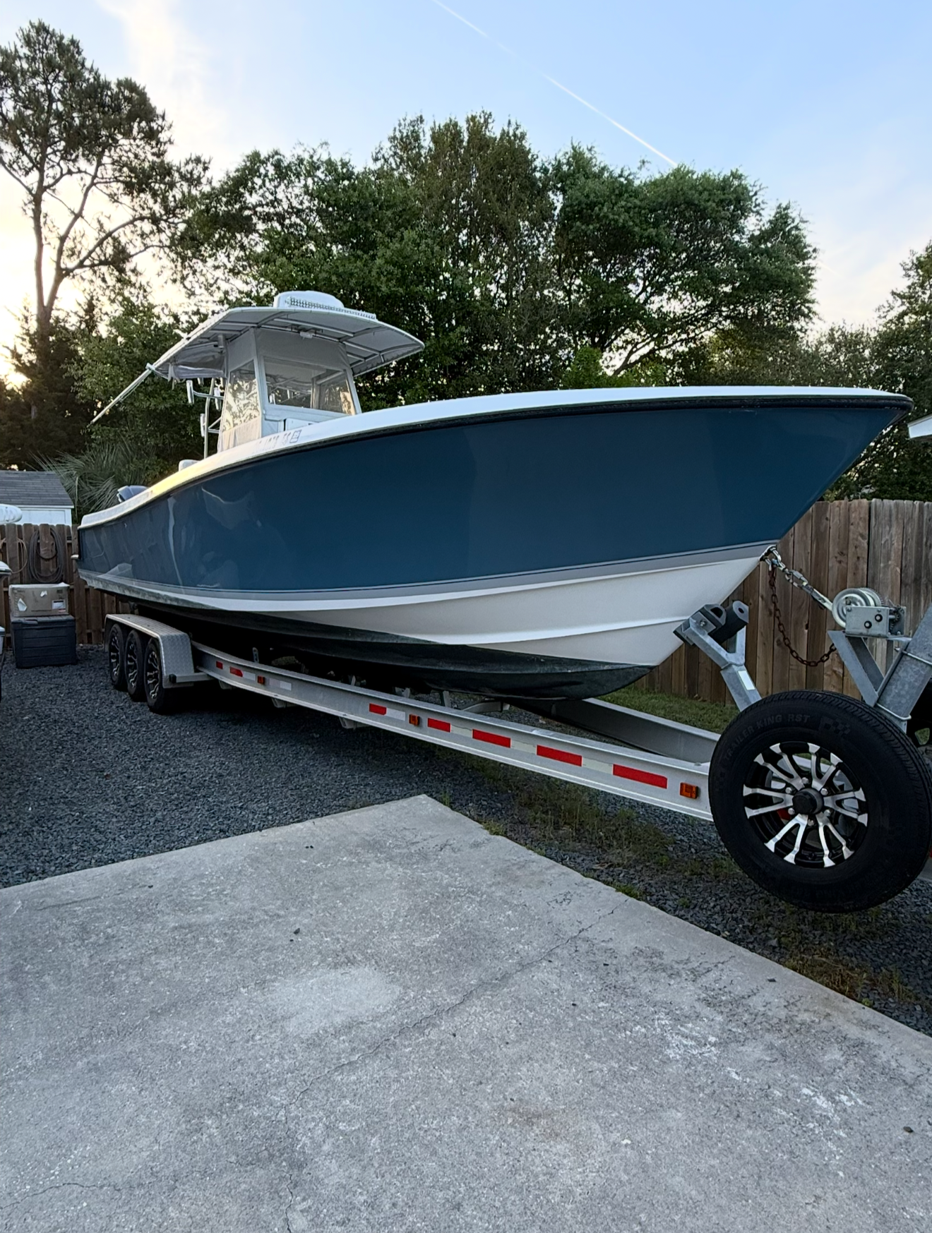 A blue and white speedboat on a trailer parked on a gravel driveway, with a wooden fence and trees in the background and a partly cloudy sky overhead.