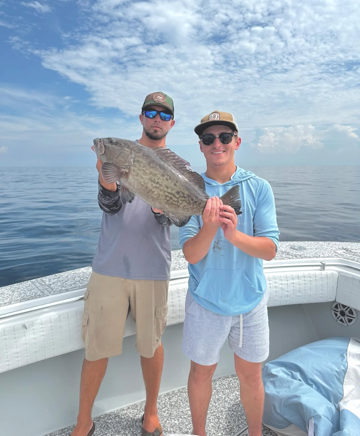Two young men on a boat holding a large fish together, with open water and a cloudy sky in the background.