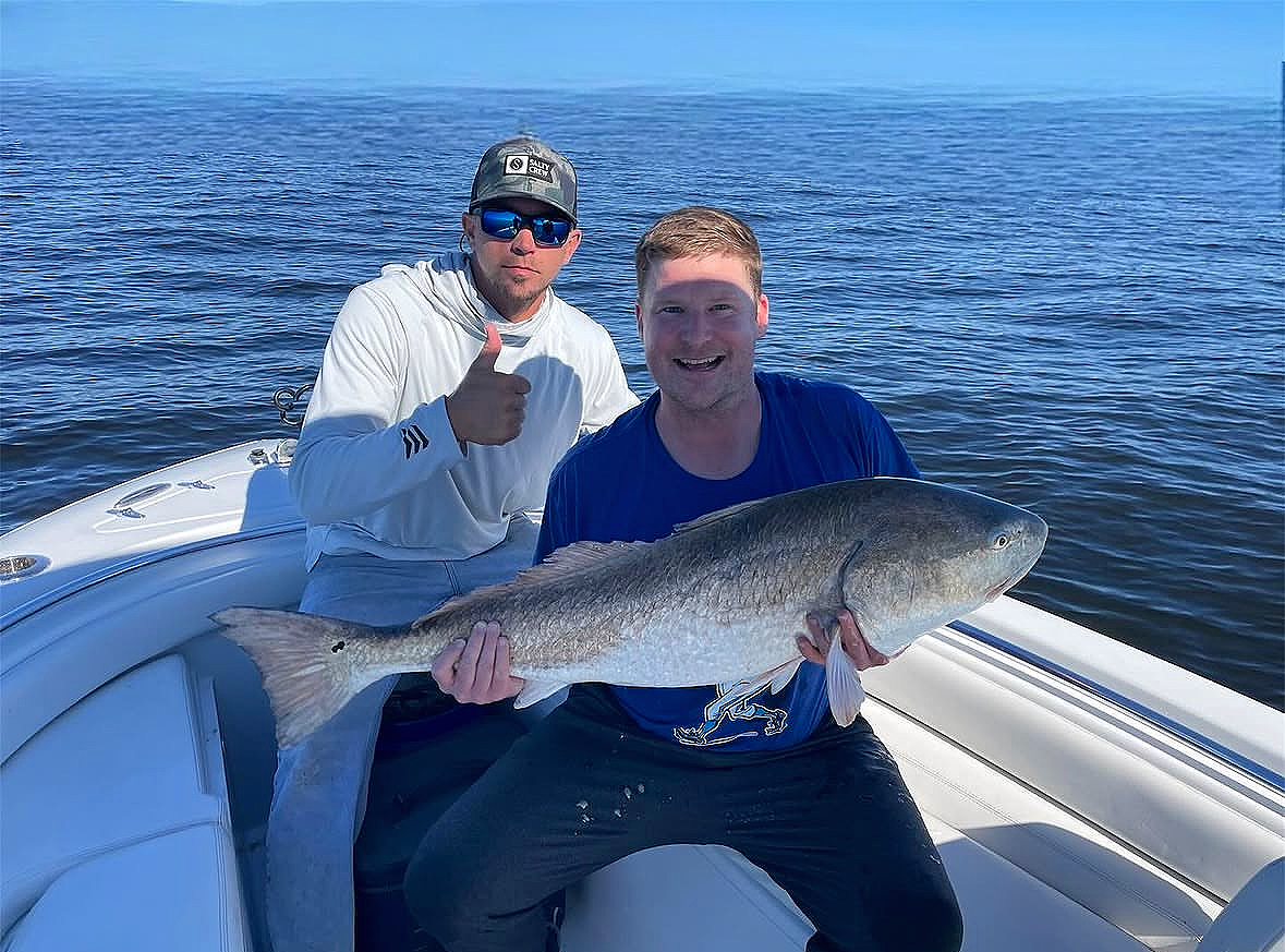 Two men on a boat holding a large fish, with one giving a thumbs up. The man in the front is smiling, wearing a blue shirt, and holding the fish with both hands. The man in the back is wearing sunglasses, a cap, and a white hoodie.