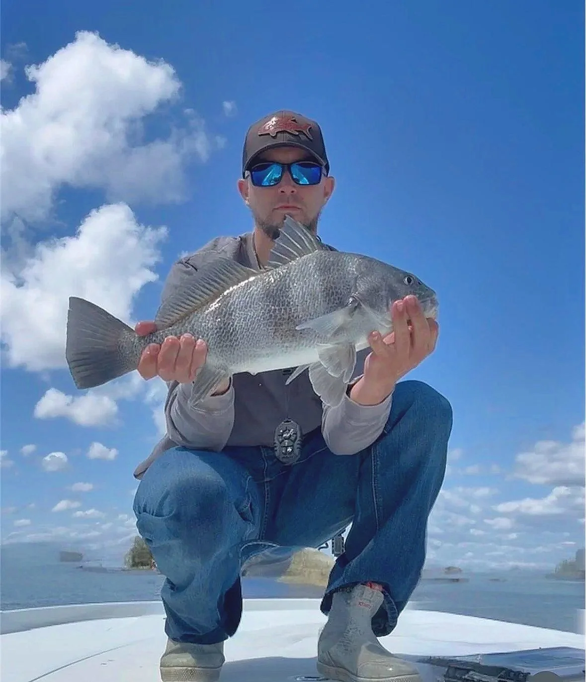 A man wearing sunglasses, a baseball cap with a fish emblem, and casual clothing, kneeling on a boat with a blue sky and clouds in the background, holding a large fish.