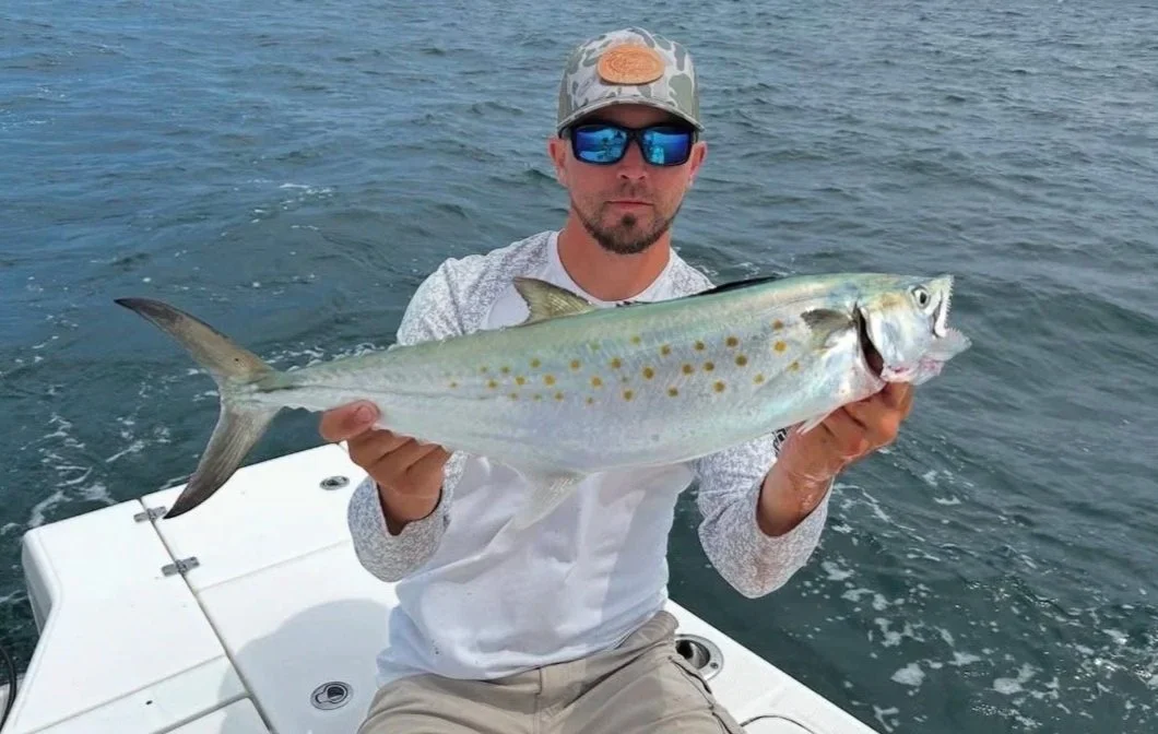 A man wearing sunglasses and a caps holding a large fish on a boat in the water.