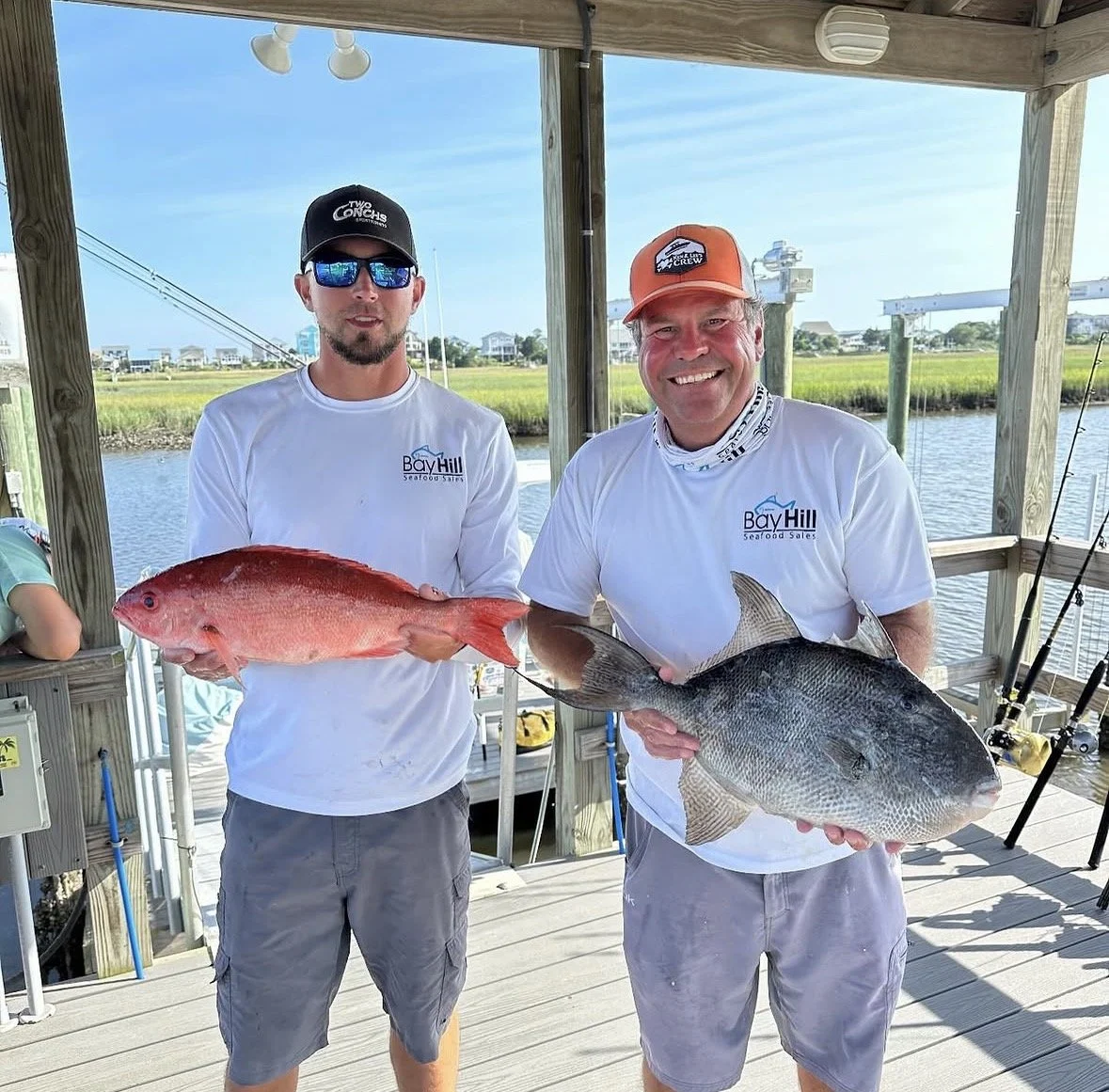 Two men standing on a wooden dock holding fish, with a waterway and marsh grass behind them. Both men are wearing white shirts with 'Bay Hill Seafood Sales' logos, and one is wearing sunglasses and a black cap while the other has an orange cap and a neck gaiter. They are smiling and show off their catch.