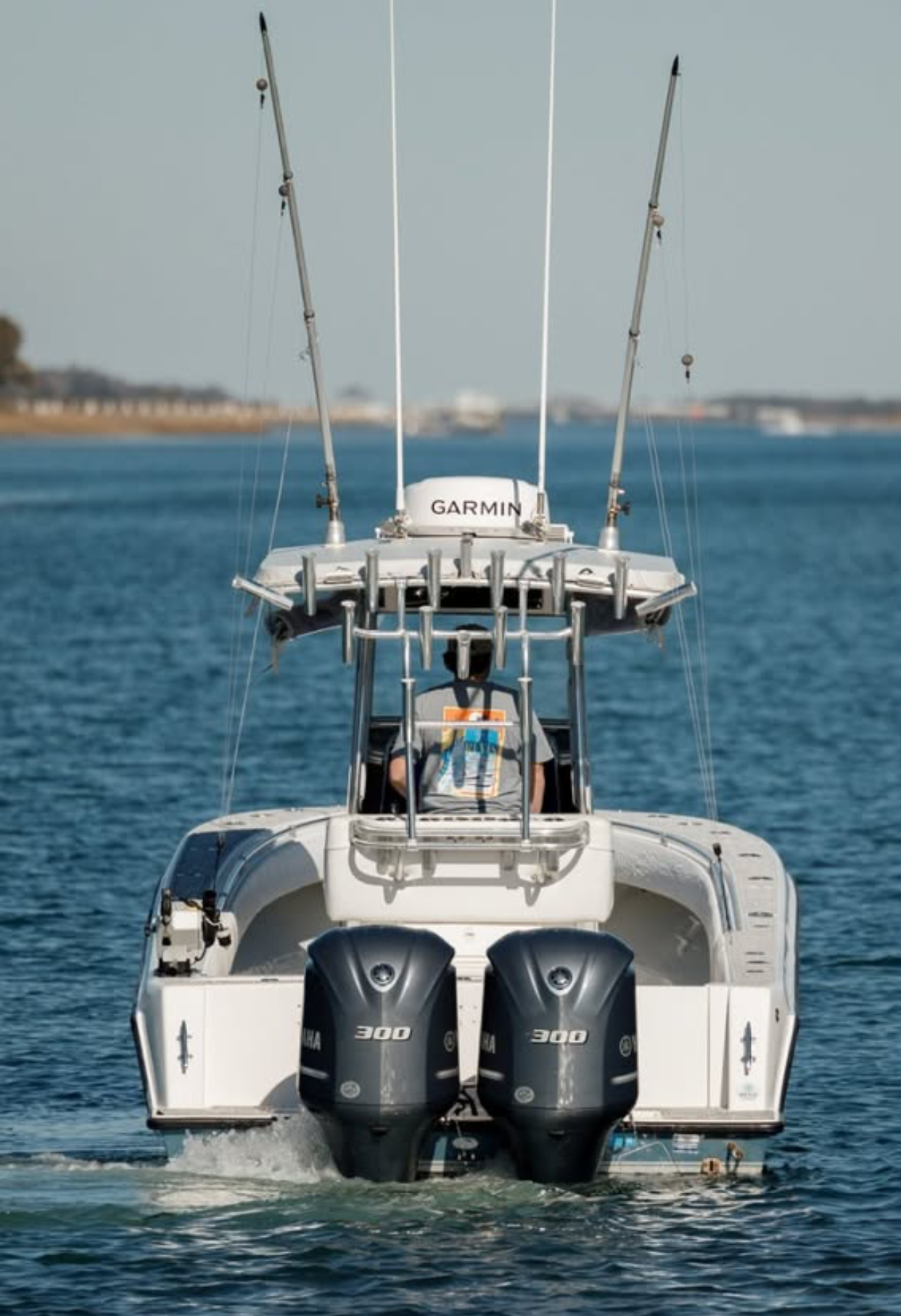 A white motor yacht on calm water with two outboard engines, two fishing rods on the upper deck, and a person in a gray shirt sitting in the back.