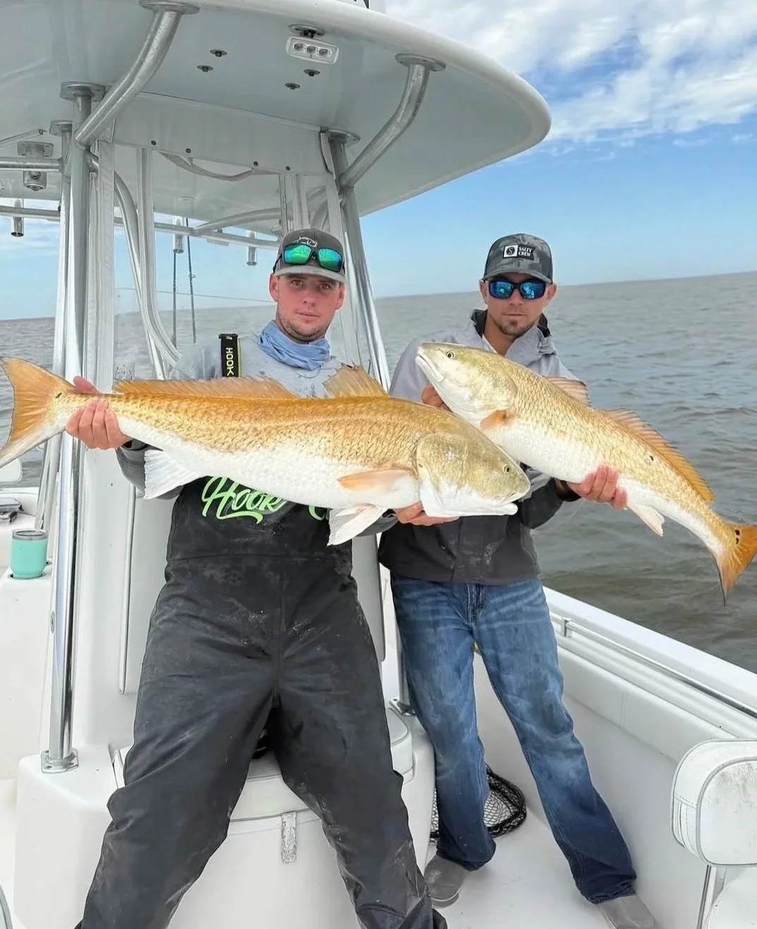 Two men on a boat holding large fish they caught, with water and sky in the background.