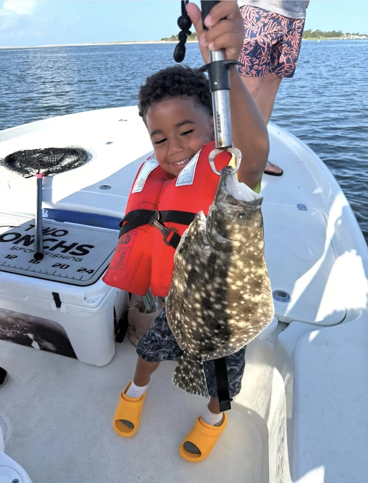 Young boy smiling on a boat, holding up a fish he caught with a fish scale. The boy is wearing a red life jacket, patterned shorts, yellow Crocs, and a gray t-shirt. The boat is on a body of water with a shoreline in the background.