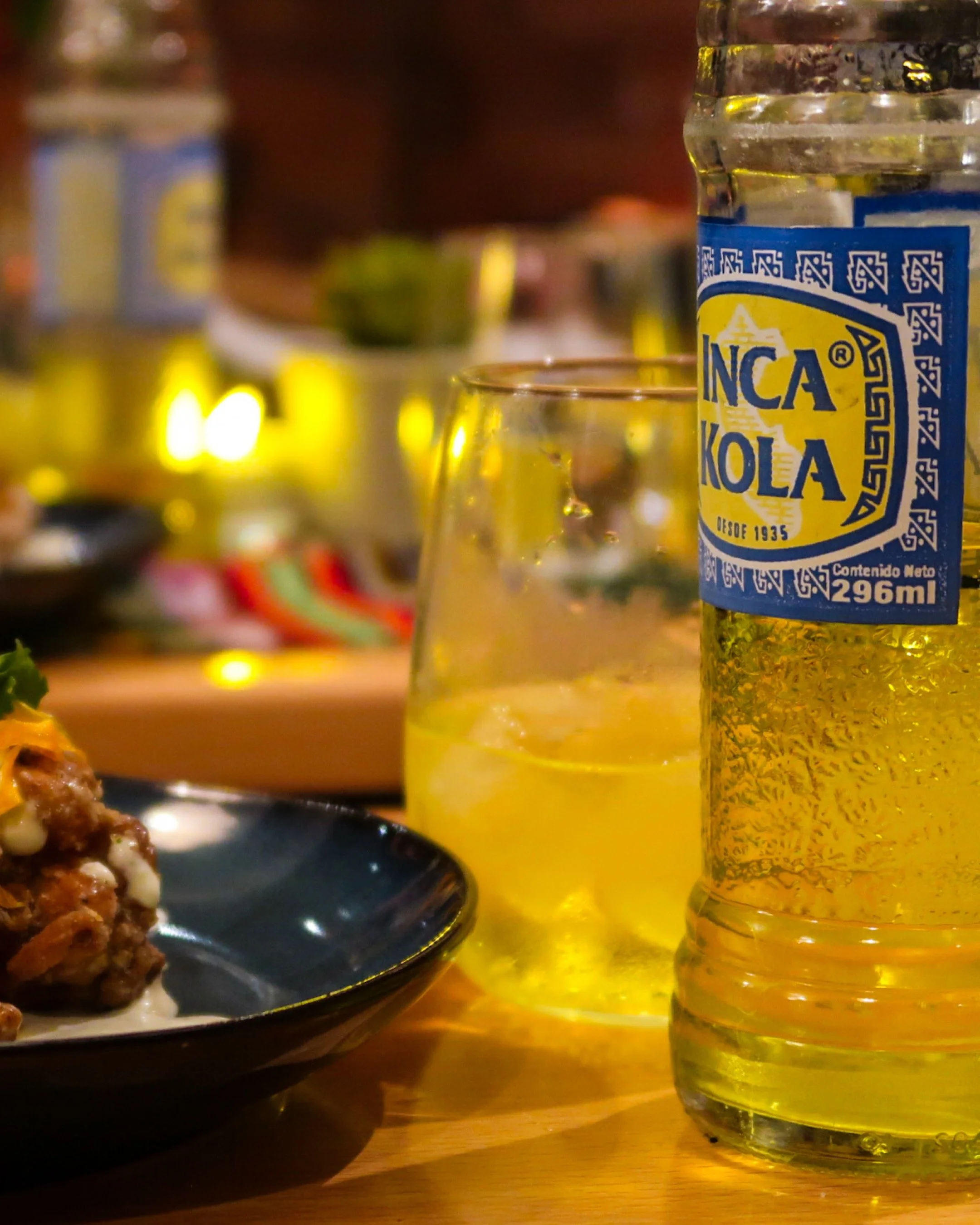 A bottle of Inca Kola soda, a glass with ice and lemon slice, and a dark bowl with food on a wooden table.