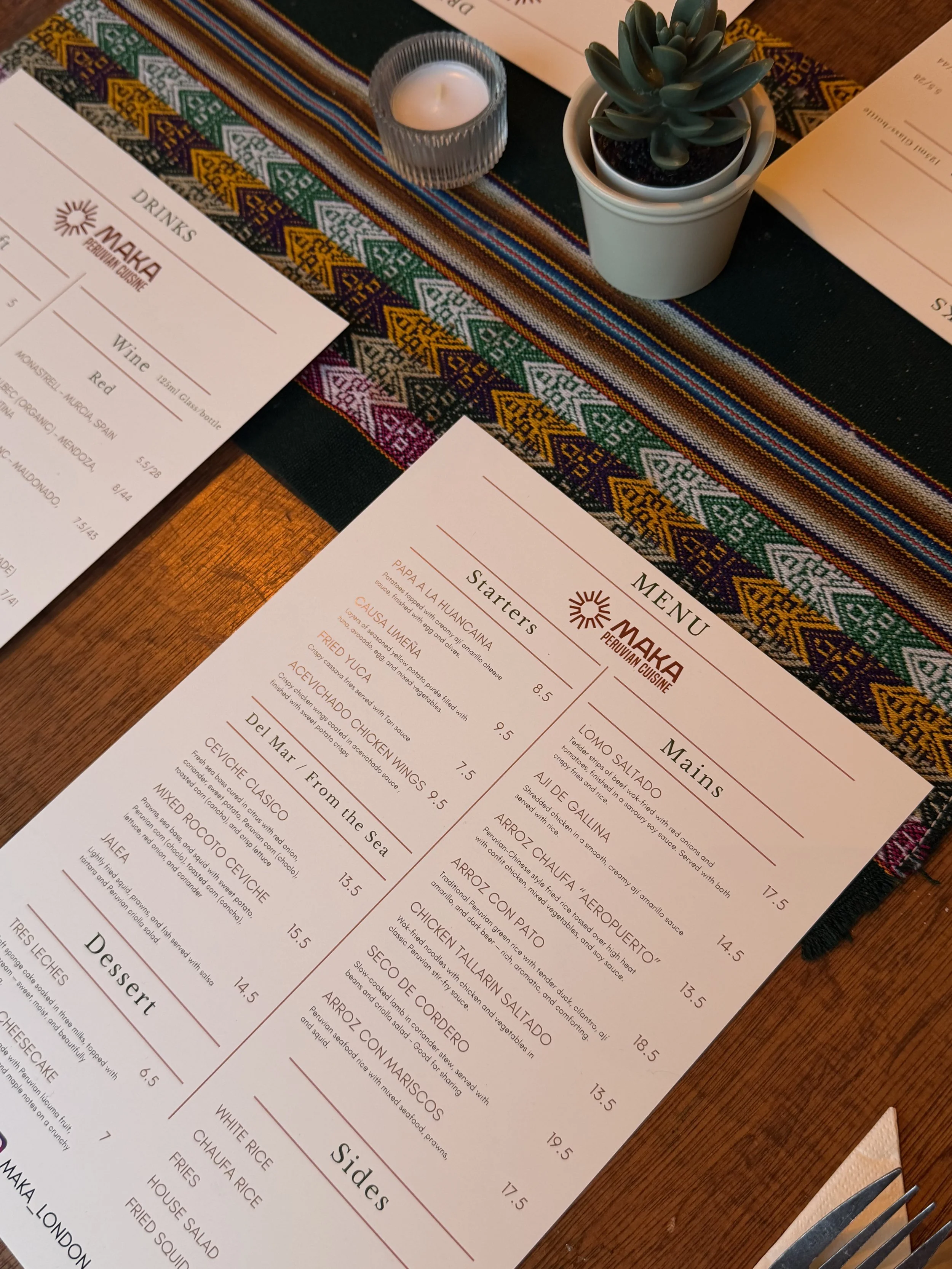 Restaurant menu on a table with a colorful textile table runner, a potted succulent, a lit candle, and cutlery.
