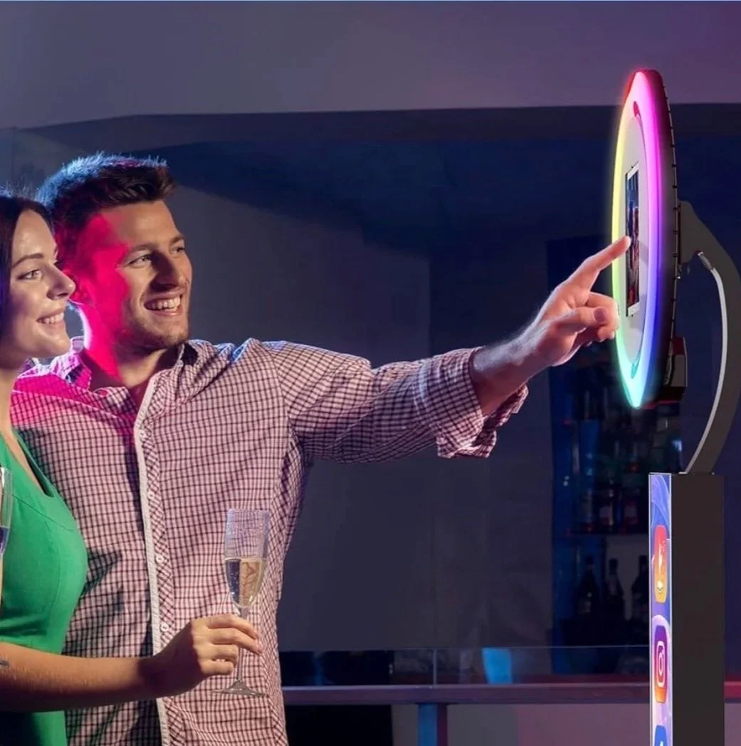 A man and woman at a party looking at a colorful, rainbow-lit digital photo frame with social media icons displayed, one person holding a glass of champagne.