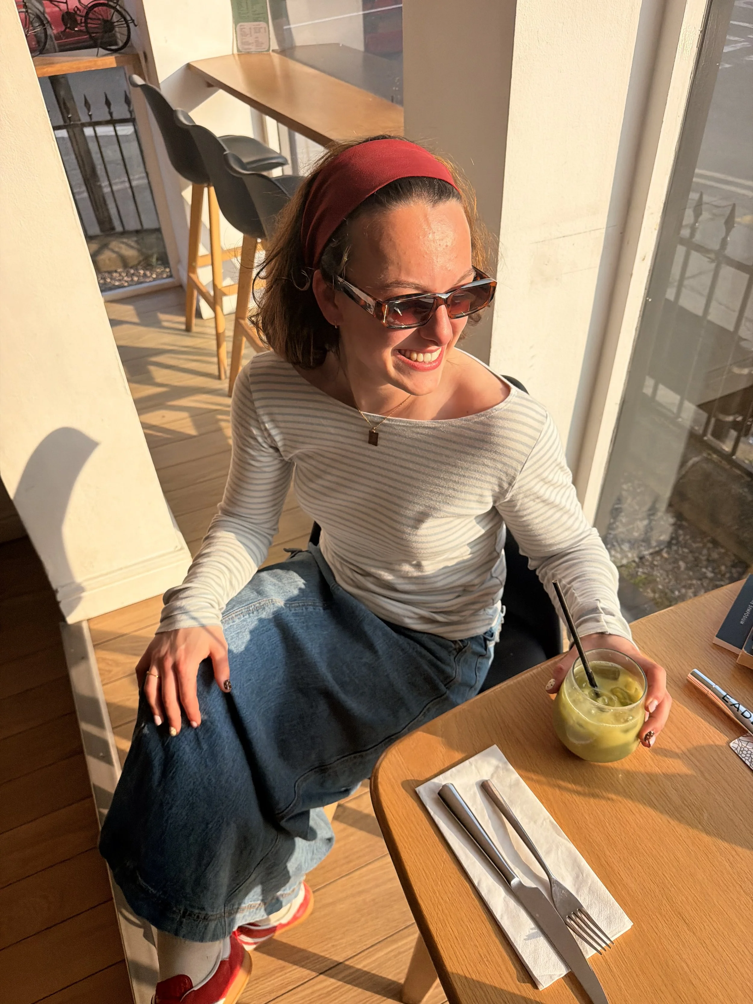 A woman with short brown hair, wearing a red headband, sunglasses, a striped long sleeve shirt, and baggy jeans, is sitting at a wooden table in a cafe, smiling and holding a glass of green drink with a straw.