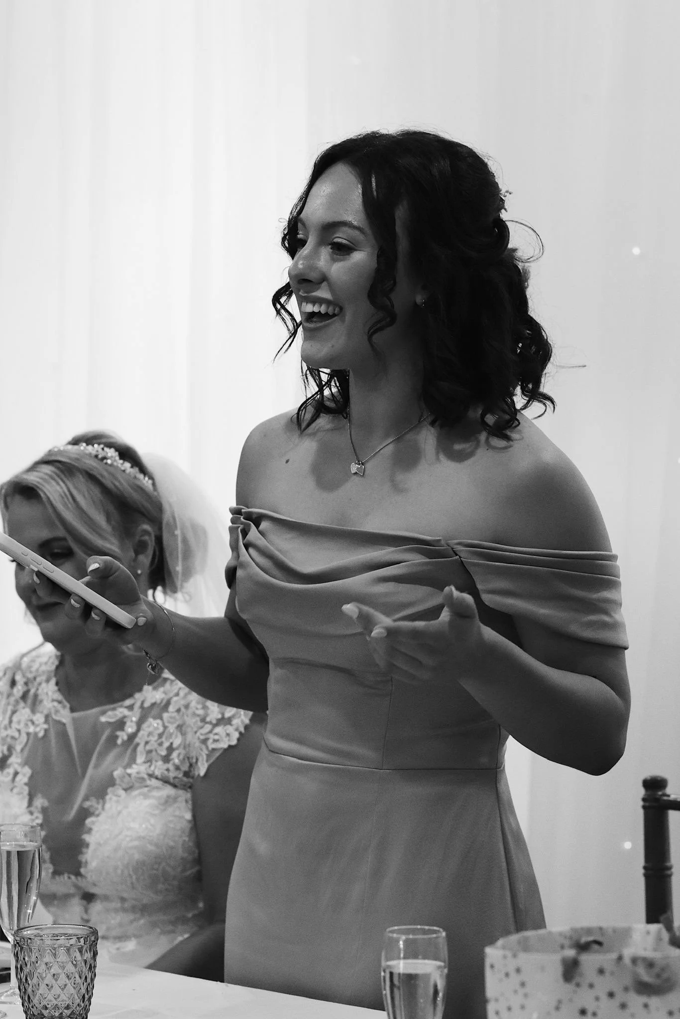 A woman in an off-the-shoulder dress is smiling and talking, holding a smartphone, with a woman in a lace dress and tiara sitting beside her.