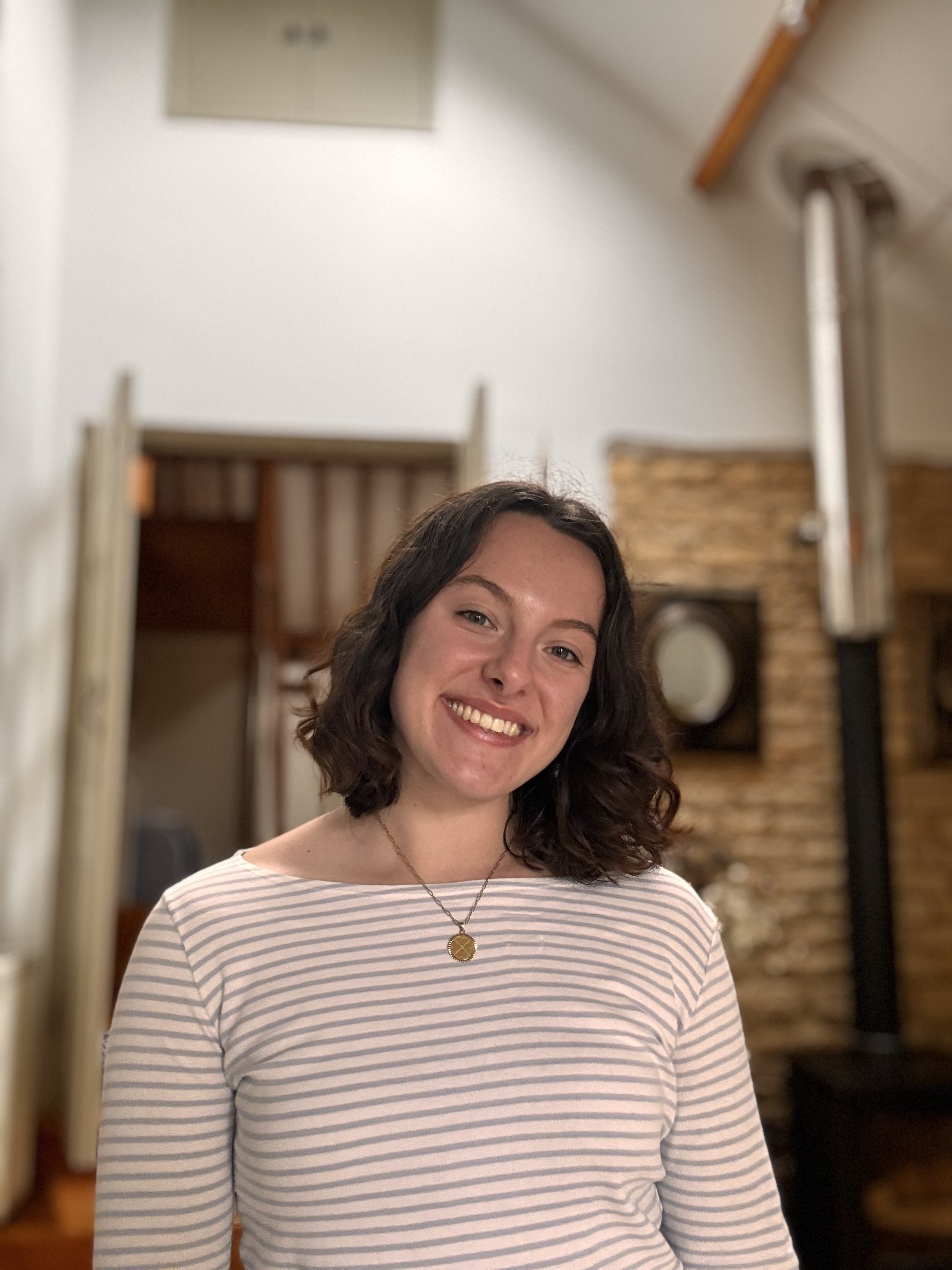 A young woman with shoulder-length dark hair smiling indoors, wearing a striped long-sleeve shirt and a necklace, with a blurred background of a room with brick wall and wooden elements.