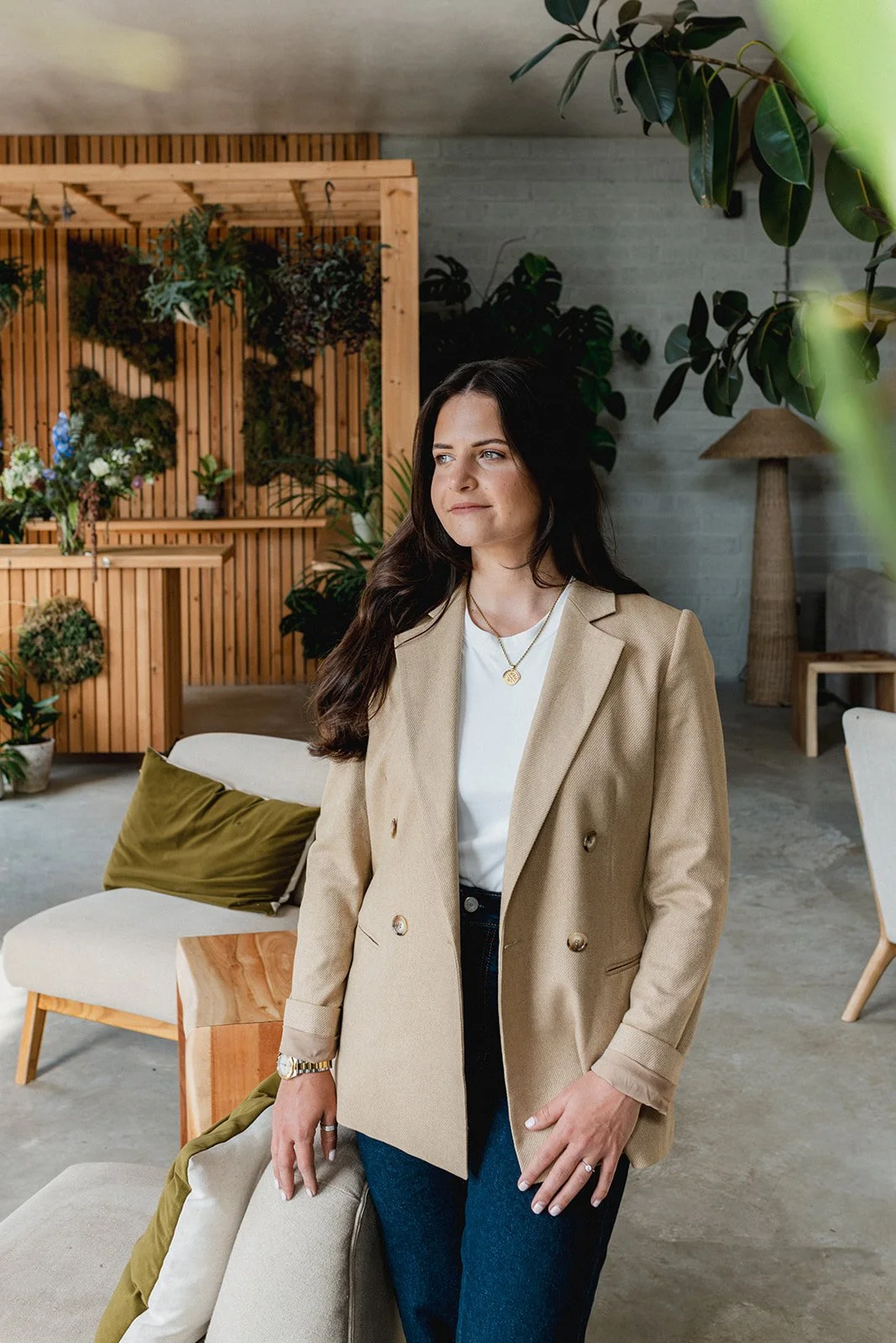 A woman with long dark hair wearing a beige blazer and white shirt standing in a modern living room with plants and wooden decor.