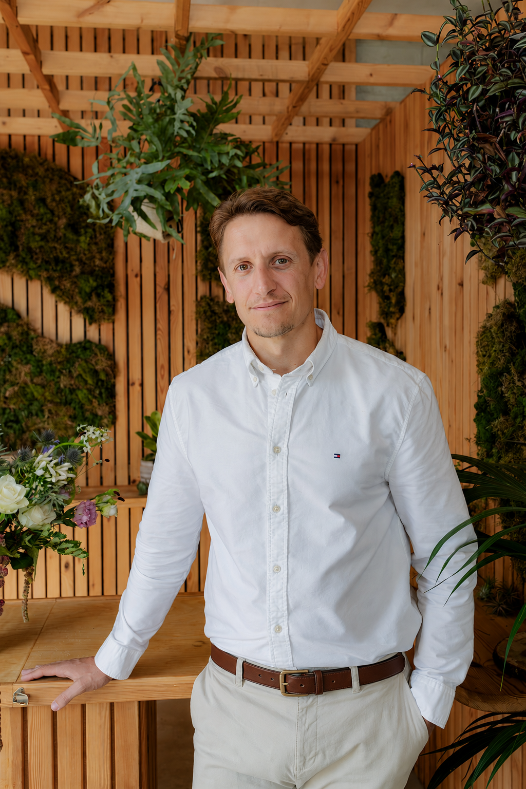 A man with brown hair and a light beard wearing a white button-down shirt and beige pants, standing in a room with wooden walls and various green plants and flowers in the background.