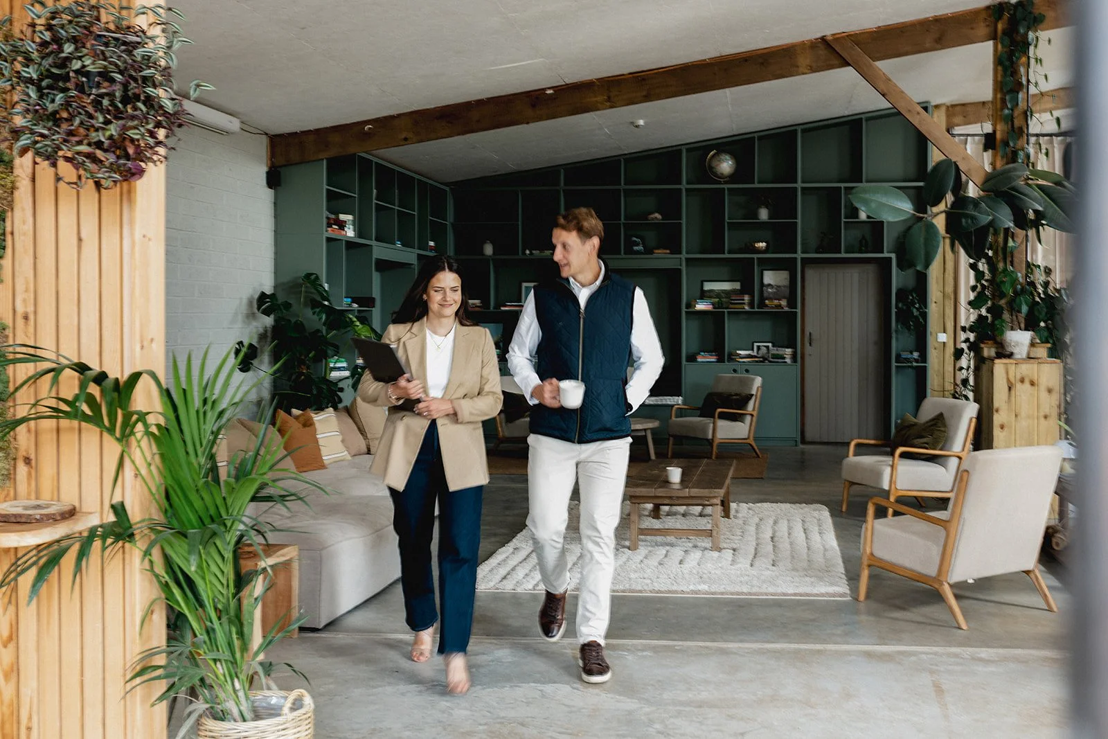 Two people walking and talking in a cozy, modern living room, with plants and wooden accents.