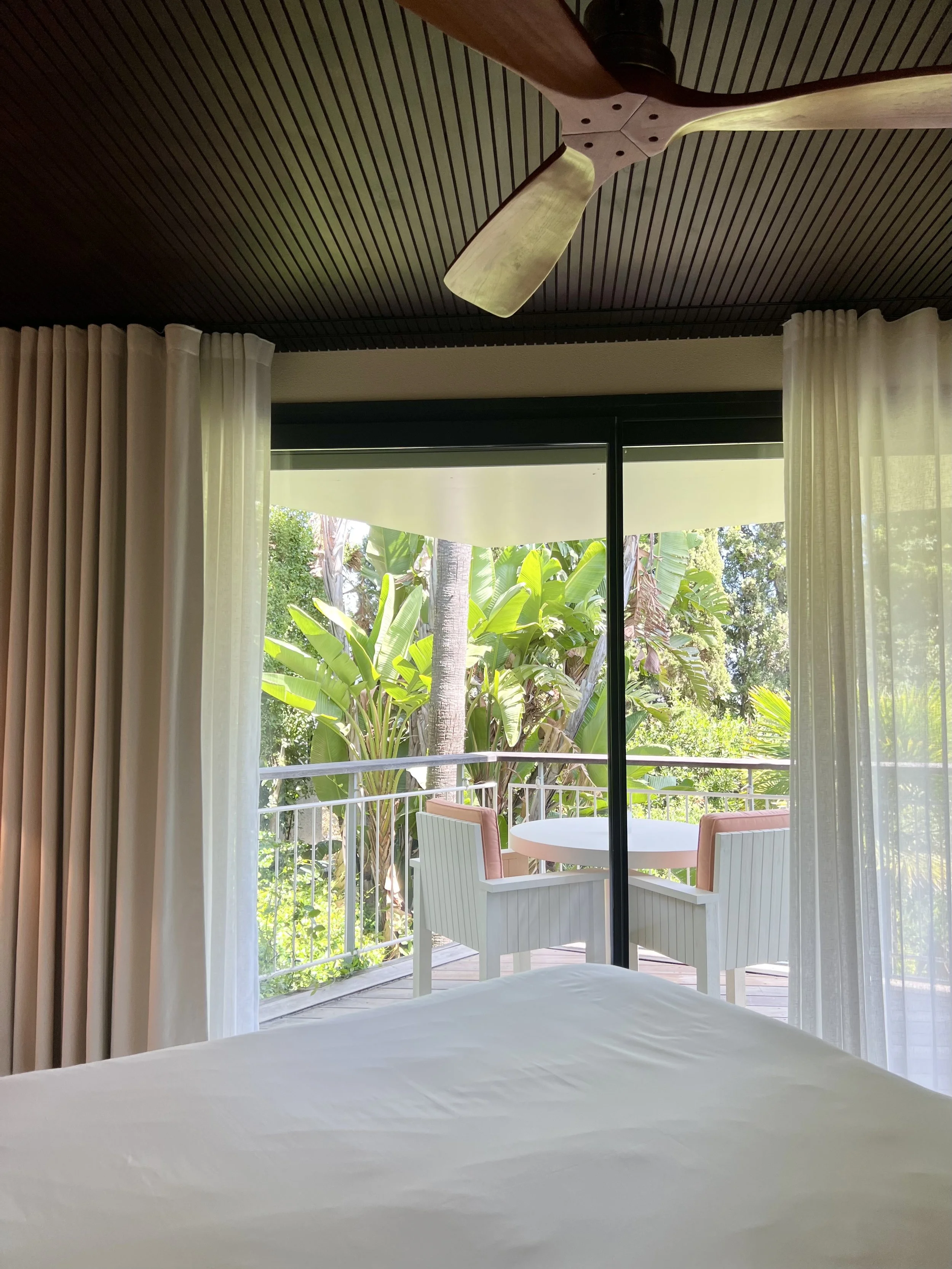 View from inside a hotel room looking out through a glass sliding door to a balcony with outdoor chairs and tropical greenery.