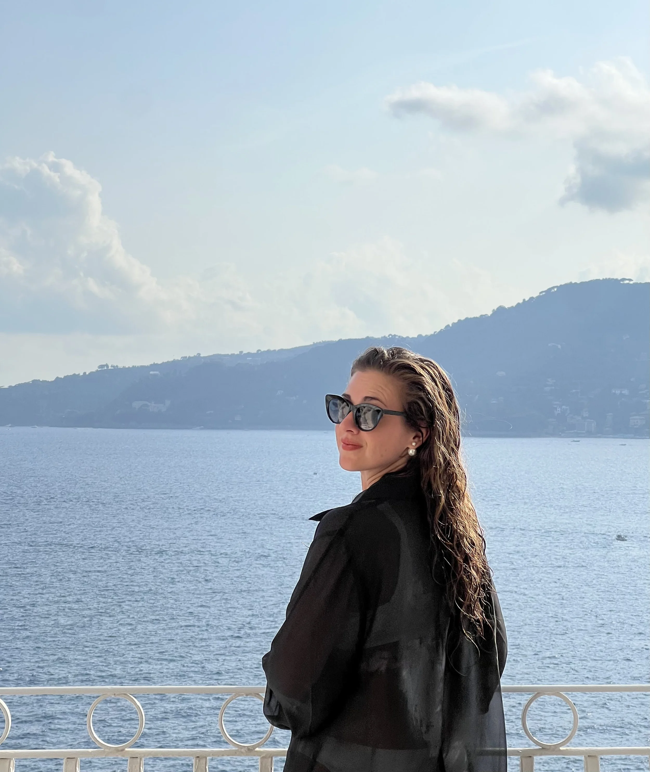 A woman with long, wet hair wearing sunglasses and earrings standing by a white railing near a body of water, with hills and a cloudy sky in the background.