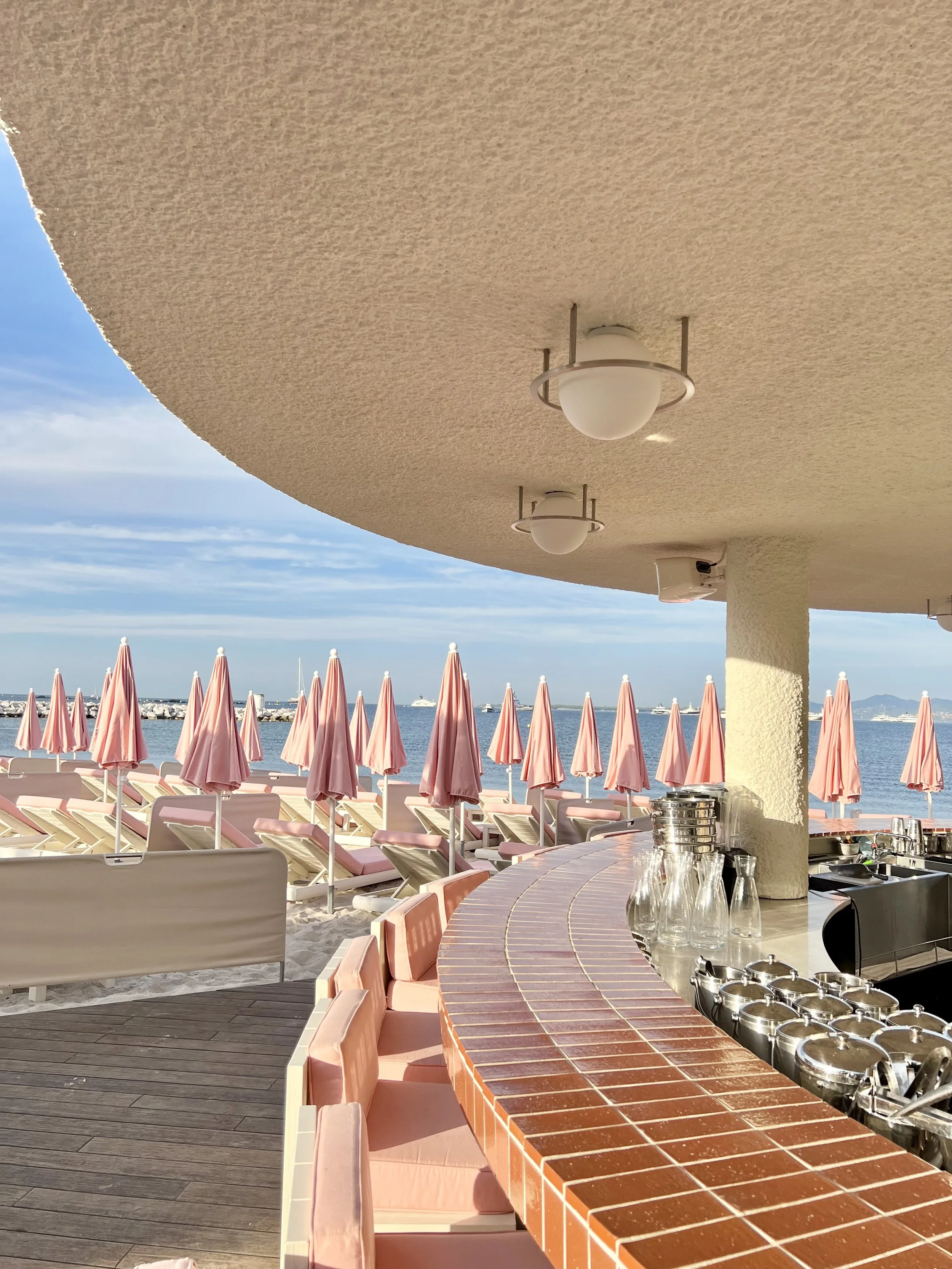 Beachside bar with pink umbrellas and lounge chairs, open seating area with pink cushions, bar counter with glassware and bottles, ocean view and boats in the distance under a partly cloudy sky.