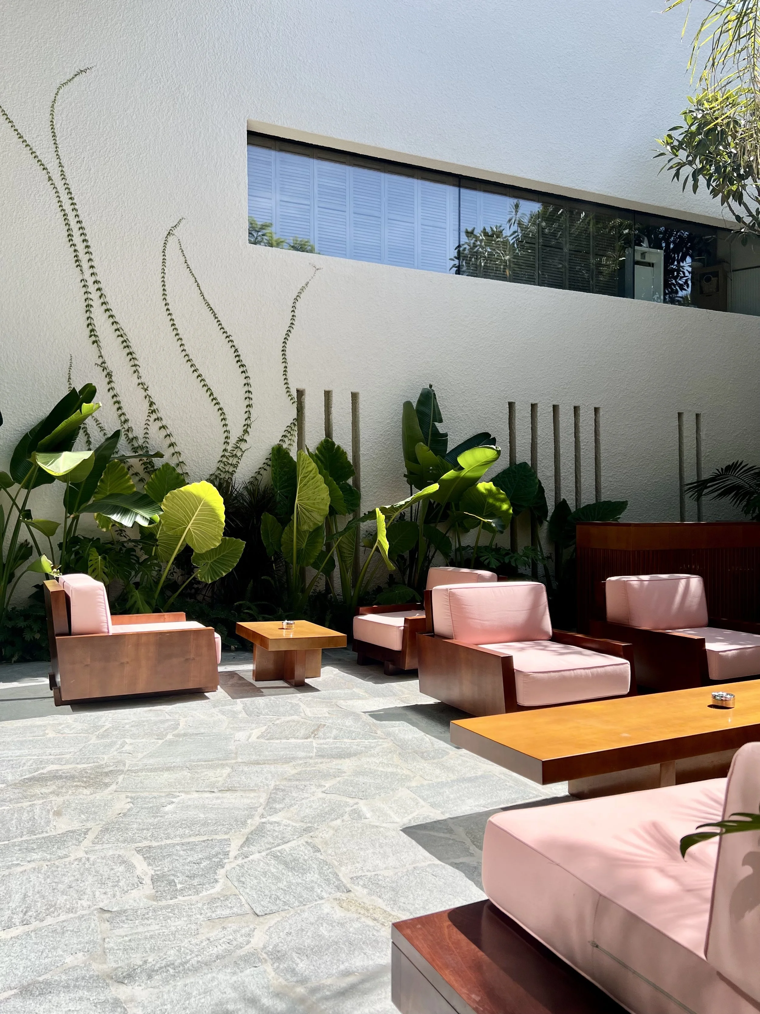 Outdoor patio area with pink cushioned wooden chairs and tables, surrounded by green tropical plants and a white wall with a window.