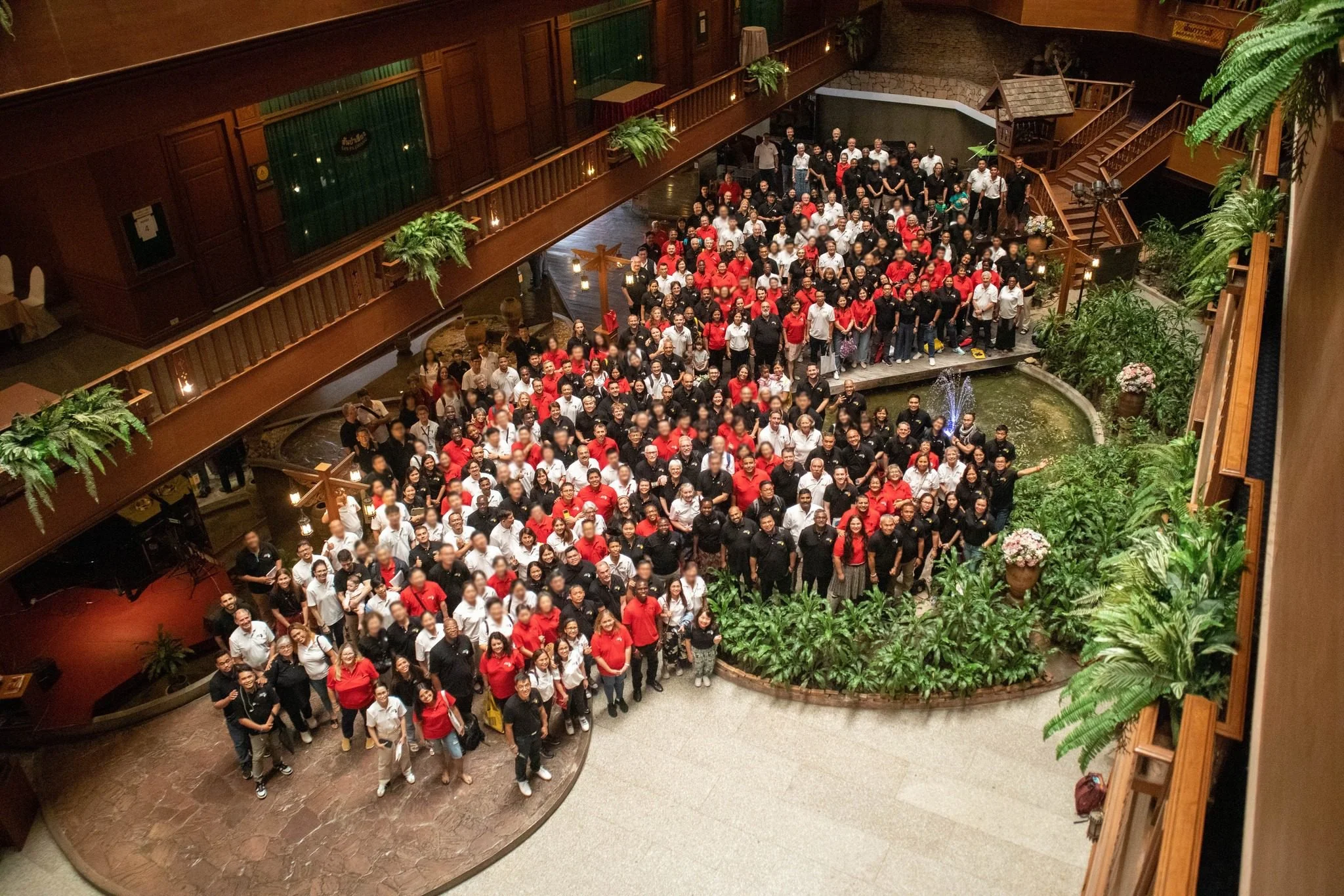 Grupo grande de personas en un evento dentro de un edificio con plantas y decoración de madera, algunas personas llevan uniformes blancos, negros o rojos.