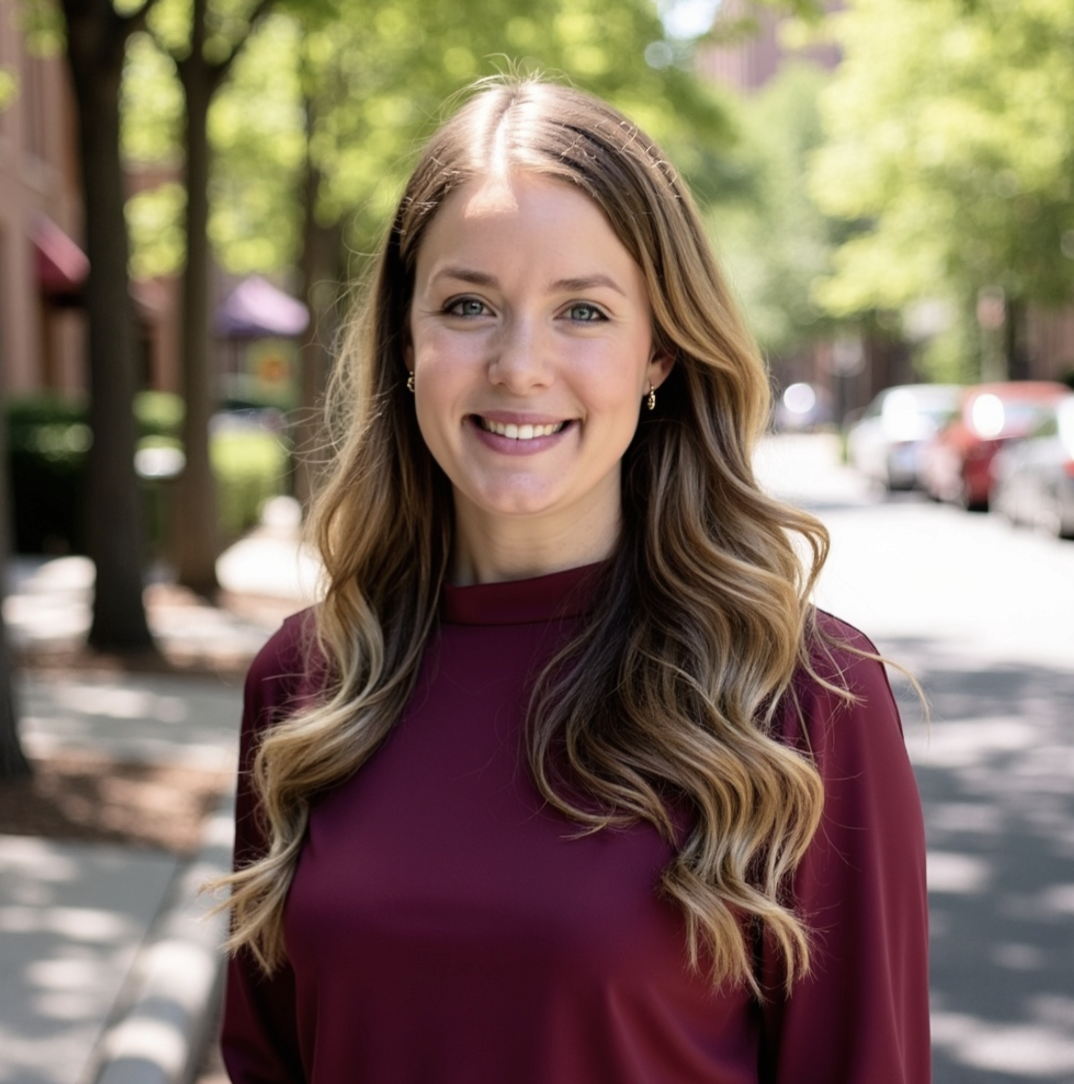 A young woman with long wavy brown hair, smiling, standing outdoors on a sunny day with trees and parked cars in the background.