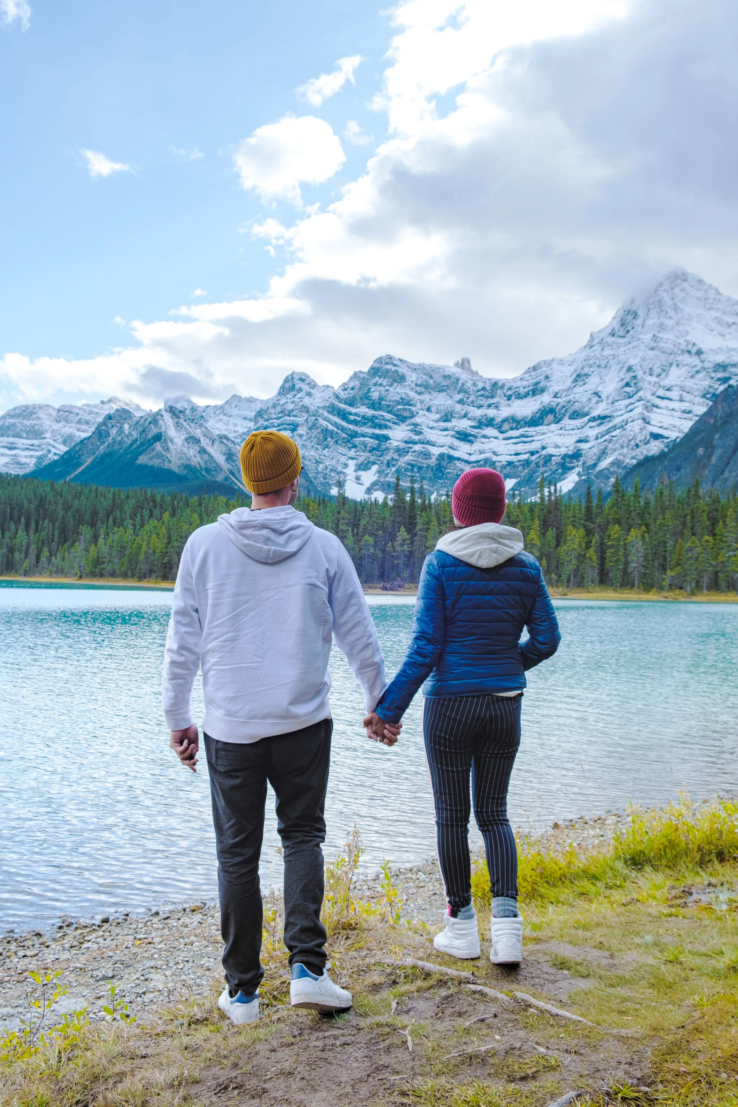 Two people, one wearing a yellow beanie and white hoodie, and the other wearing a red beanie and blue jacket, hold hands and look at a mountain lake with snow-capped mountains and a cloudy sky in the background.