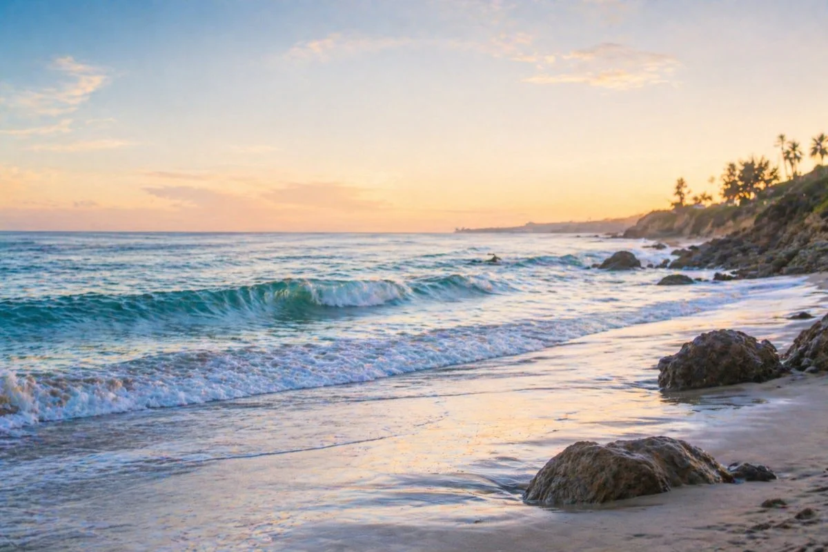 Sunset over the ocean with waves crashing onto a sandy beach, rocks on the shore, and palm trees in the distance.