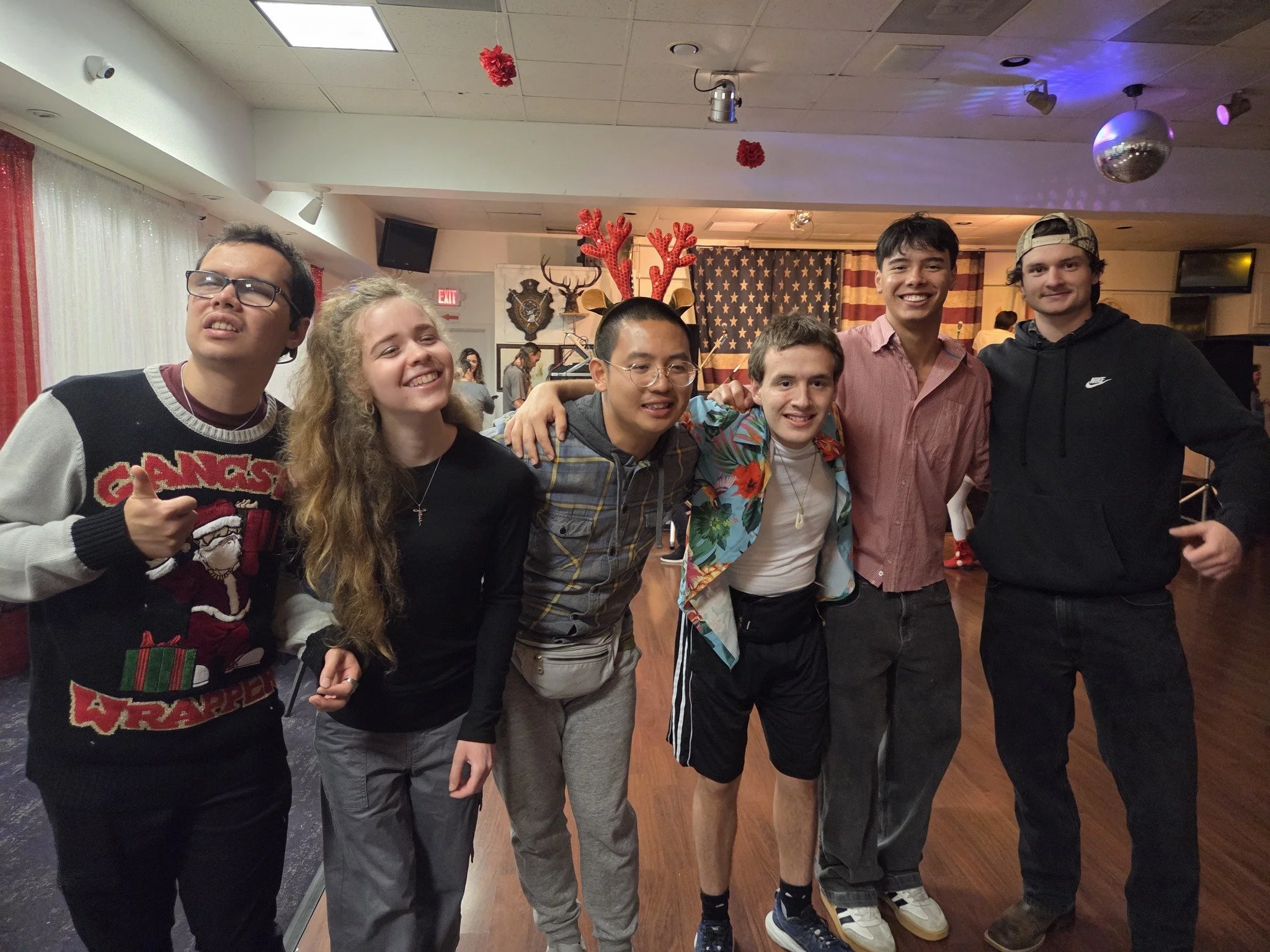 Seven young people are standing together in a room decorated with patriotic American flags and red decorations. They are smiling and posing for the photo, some with arms around each other.