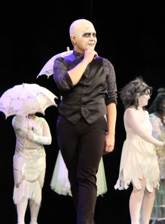 People in costume, with the central figure dressed as a mime or actor with pale makeup and dark eye makeup, standing on stage during a performance.