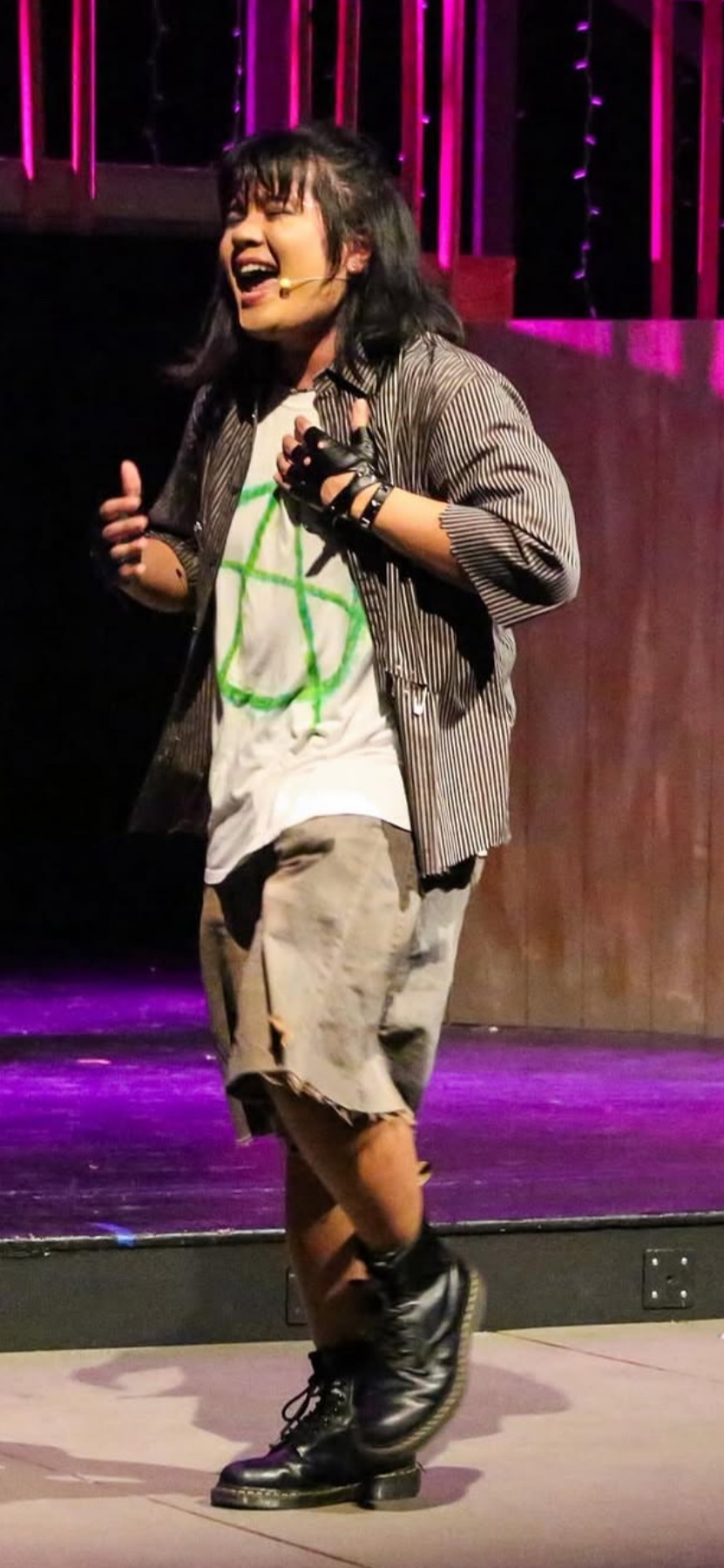 A person on stage wearing combat boots, a striped shirt over a white t-shirt with a green peace symbol, and beige shorts. They are smiling and appear to be performing or speaking into a headset microphone, with pink stage lights in the background.