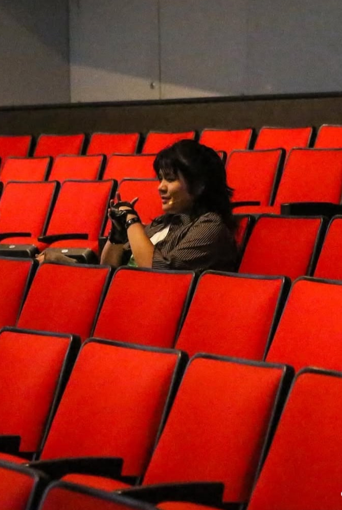 A woman sitting alone in a mostly empty theater or auditorium with red seats, looking at her phone.