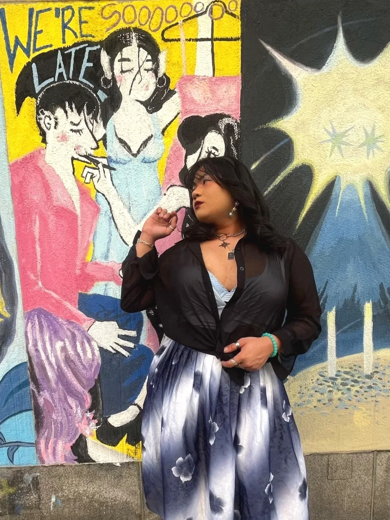 A woman with black hair wearing a black top and a flowy skirt stands in front of a colorful mural of women, one smoking, on a yellow, black, and pink background.