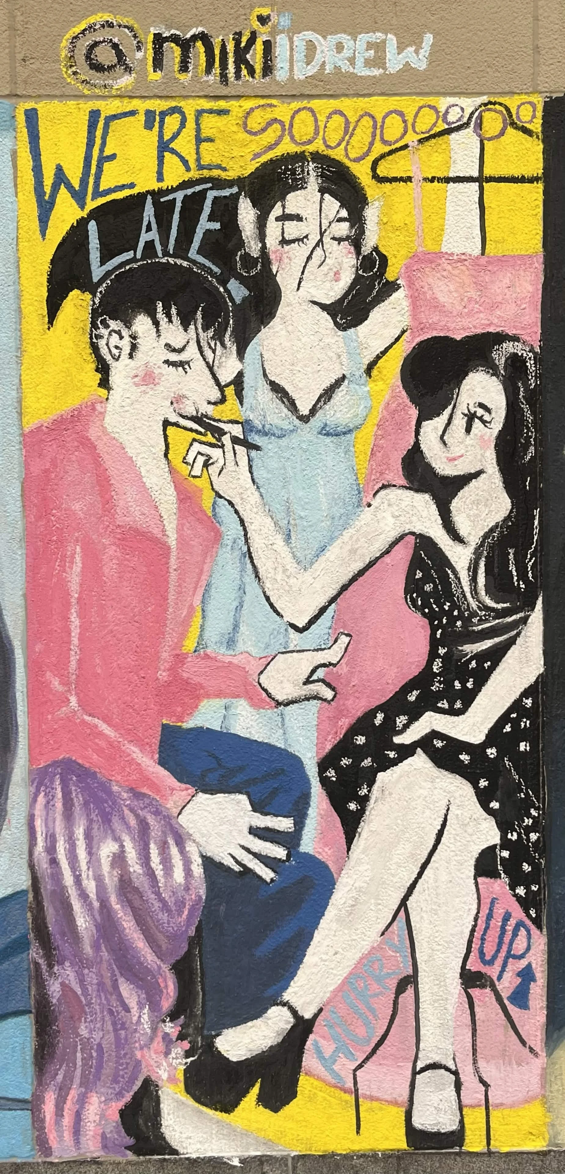 A colorful mural painting of three women gathered together. The background features bright yellow with graffiti-like text that reads 'WE'RE LATE' and some additional text at the top. One woman has short dark hair with closed eyes, wearing a pink jacket and striped skirt. Another woman has black hair in pigtails, wearing a light blue top, holding a cigarette or pen to her lips. The third woman has long black hair, wearing a black dress with a polka dot pattern, sitting on a pink chair, smiling with her eyes closed. The scene appears lively and playful.