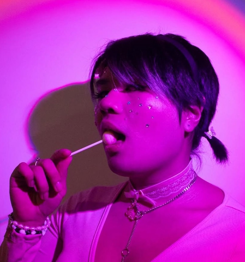 Person with dark hair adorned with sparkly embellishments on face, wearing layered necklaces and lace choker, in a pink and purple-lit setting, holding a lollipop, with a playful expression.