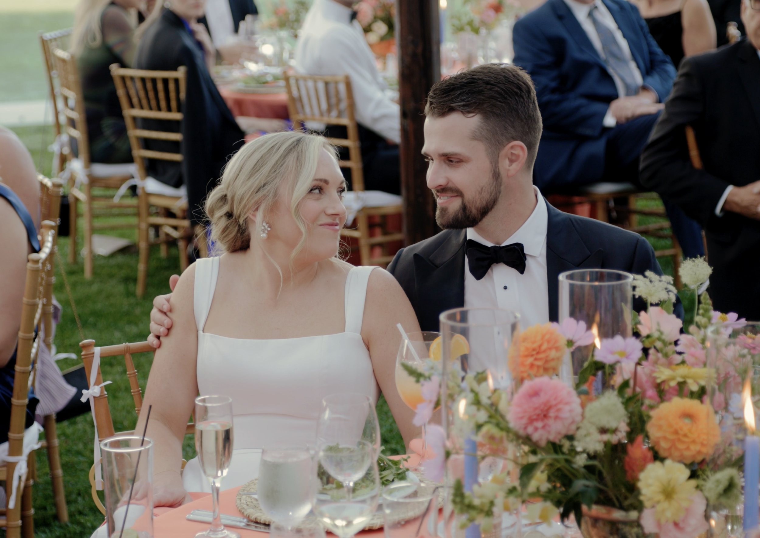 A bride and groom sitting at a wedding reception table decorated with colorful flowers, glasses, and candles, outdoors during daytime.