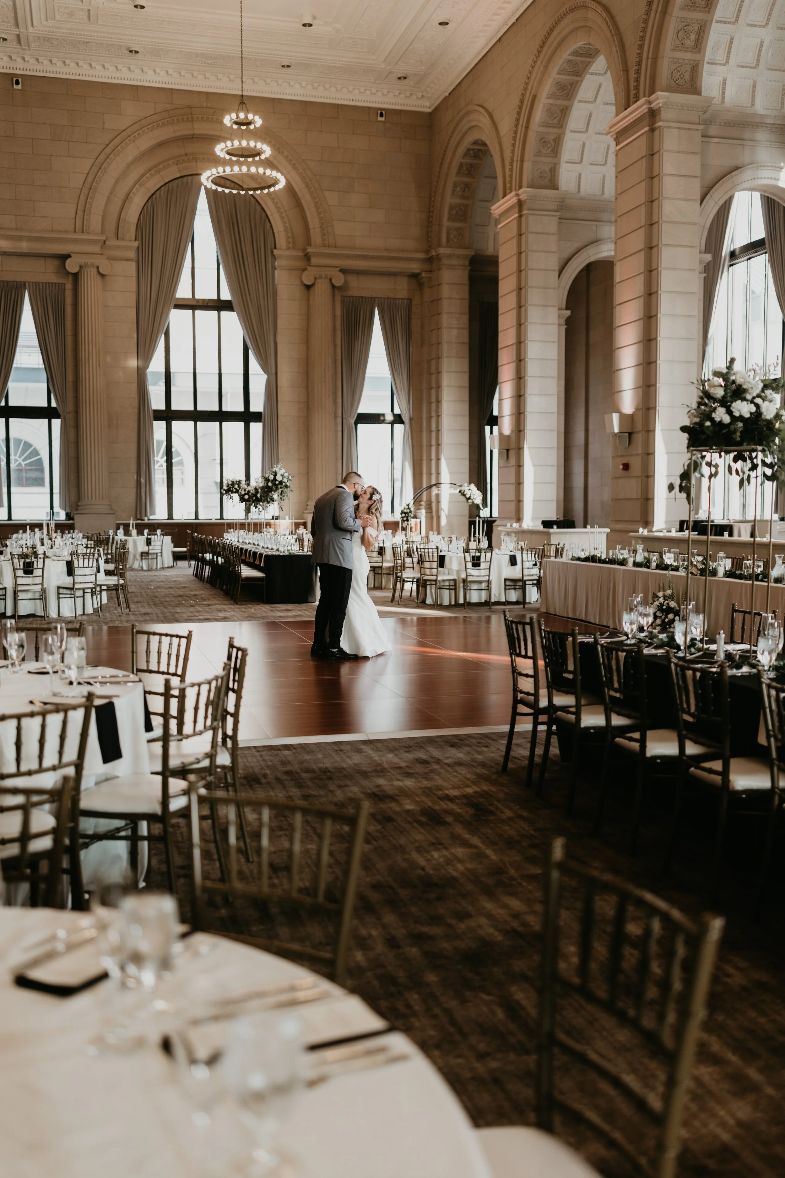 A bride and groom sharing a kiss in a large, elegant banquet hall decorated for a wedding reception, with tables set for guests and tall windows with drapes.