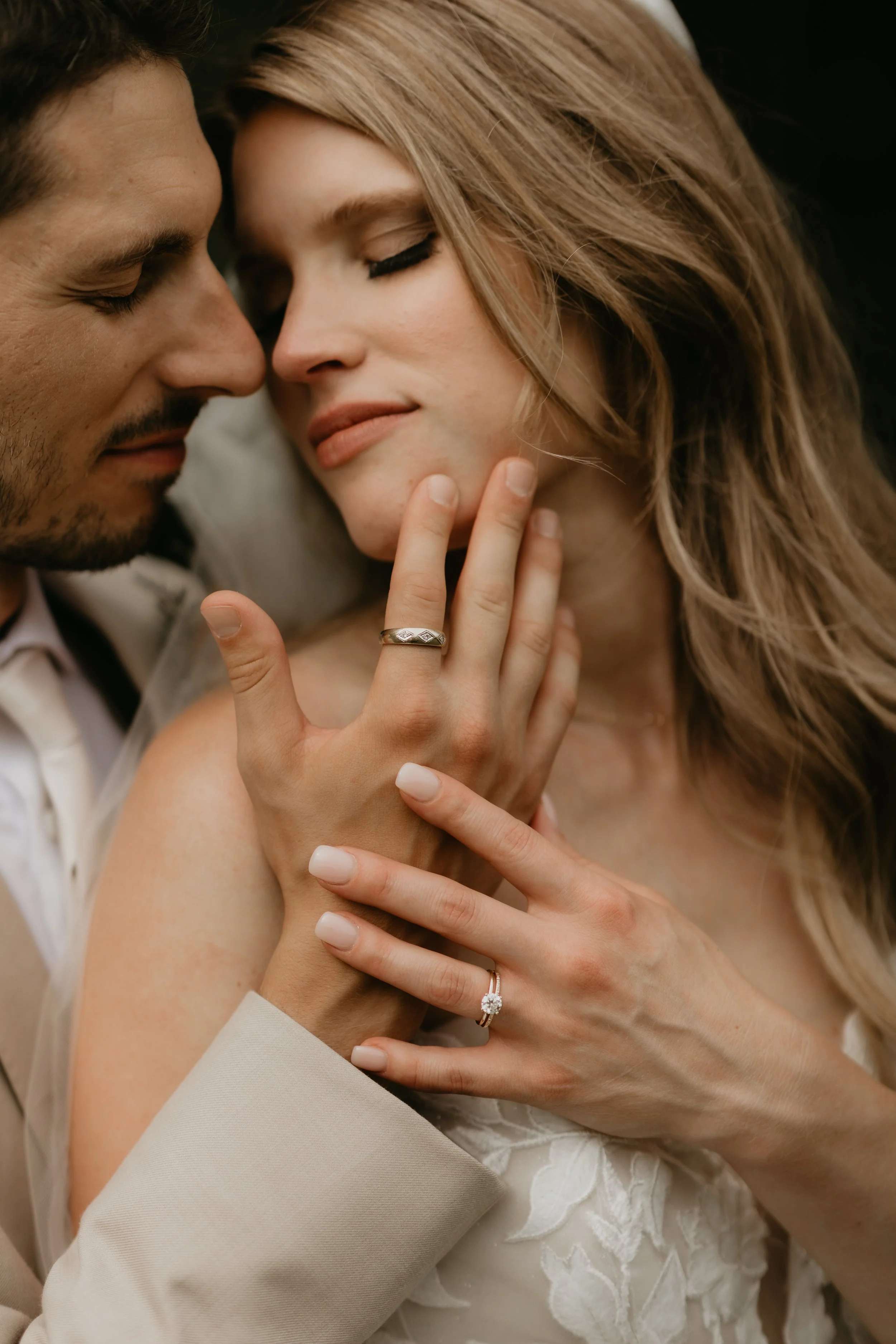 Close-up of a bride and groom in a tender moment, their foreheads touching and eyes closed, with the bride gently holding the groom's face. The bride has long, wavy hair and an engagement ring on her finger, and wearing a white lace dress. The groom has dark hair and facial hair and is dressed in a white shirt and beige suit.