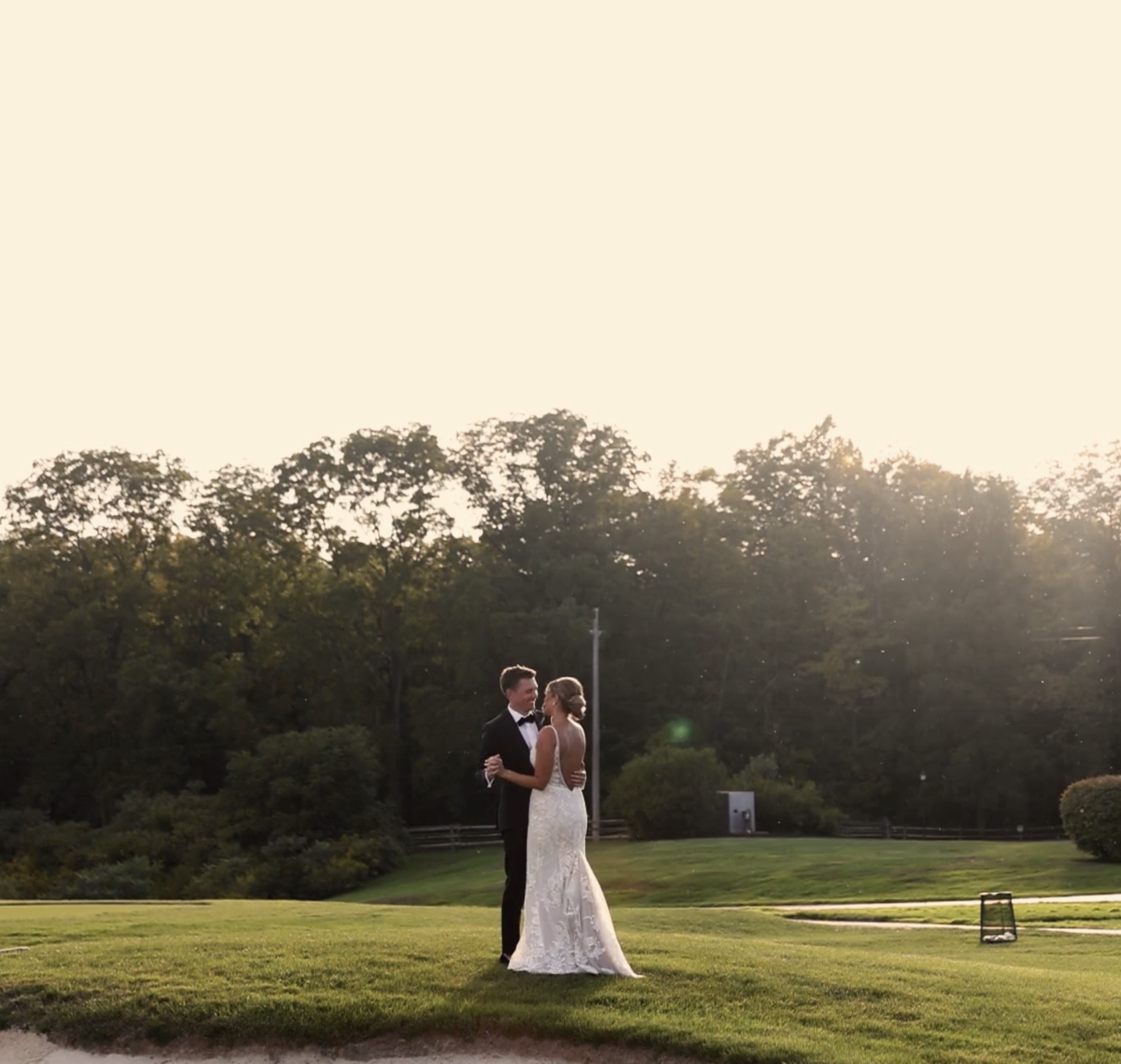 A bride and groom dancing outdoors on a grassy field during sunset, with trees in the background.