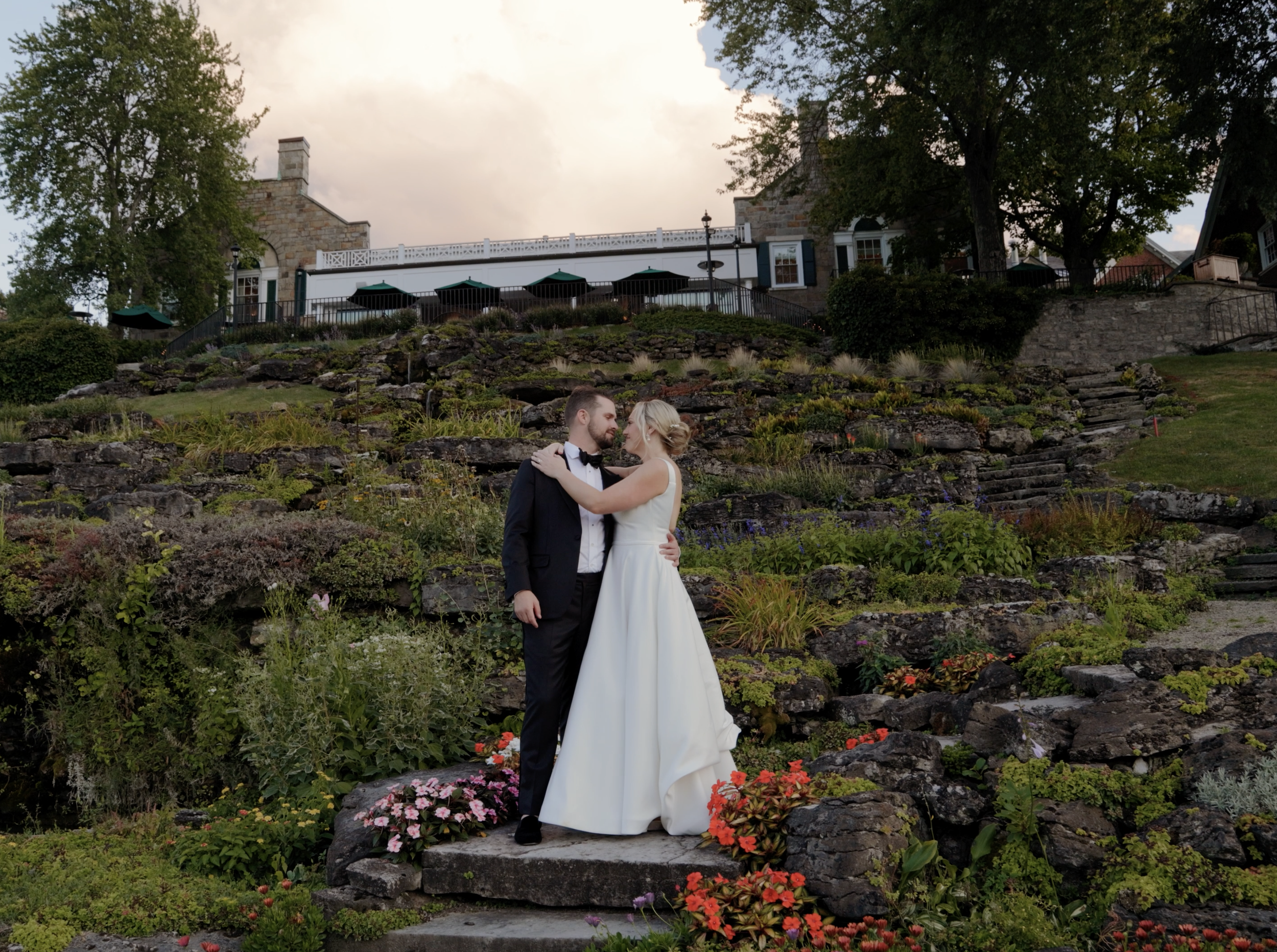 A newlywed couple in wedding attire dancing on a stone stairway, surrounded by colorful flowers, with a large house, trees, and a patio with umbrellas in the background.