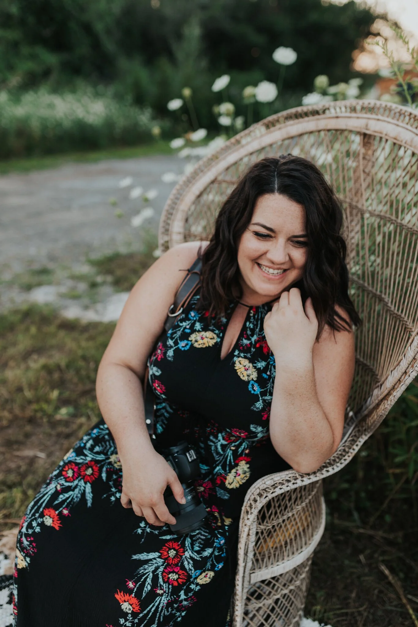 Smiling woman with dark wavy hair wearing a black dress with colorful floral embroidery, sitting in a wicker chair outdoors with greenery and white flowers in the background, holding a camera in her lap.