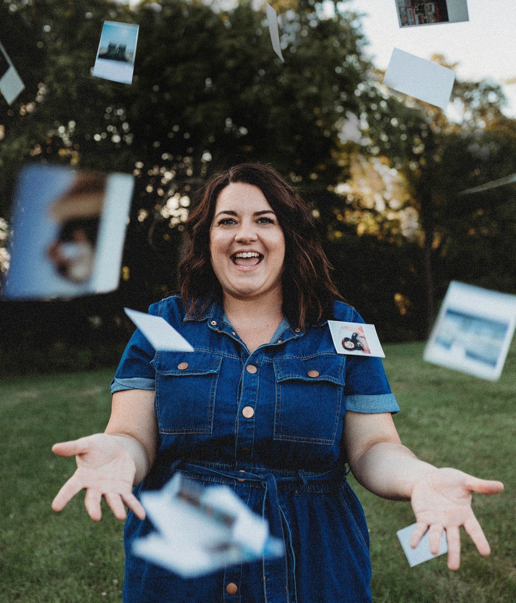 A woman with dark, wavy hair smiling, wearing a denim jacket, standing outdoors in a park-like setting with trees in the background. Photographs and possibly Polaroid pictures are floating around her, some close to the camera and others further away, giving a sense of motion or floating.