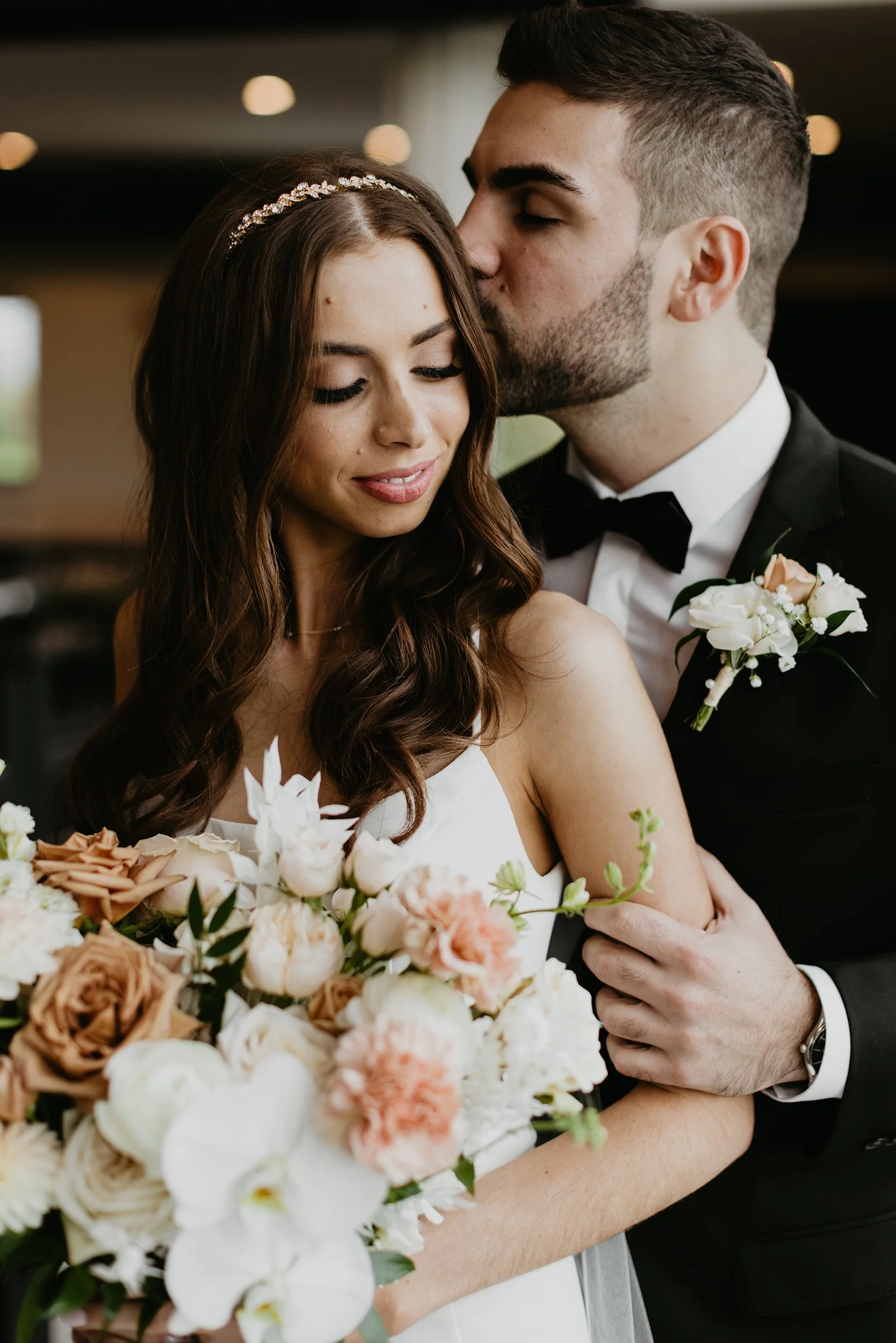 A newlywed couple, a woman with long brown hair and a man with dark hair, sharing a tender moment at their wedding. The woman is holding a bouquet of pastel flowers, and the man, dressed in a tuxedo with a boutonniere, is kissing her on the forehead.