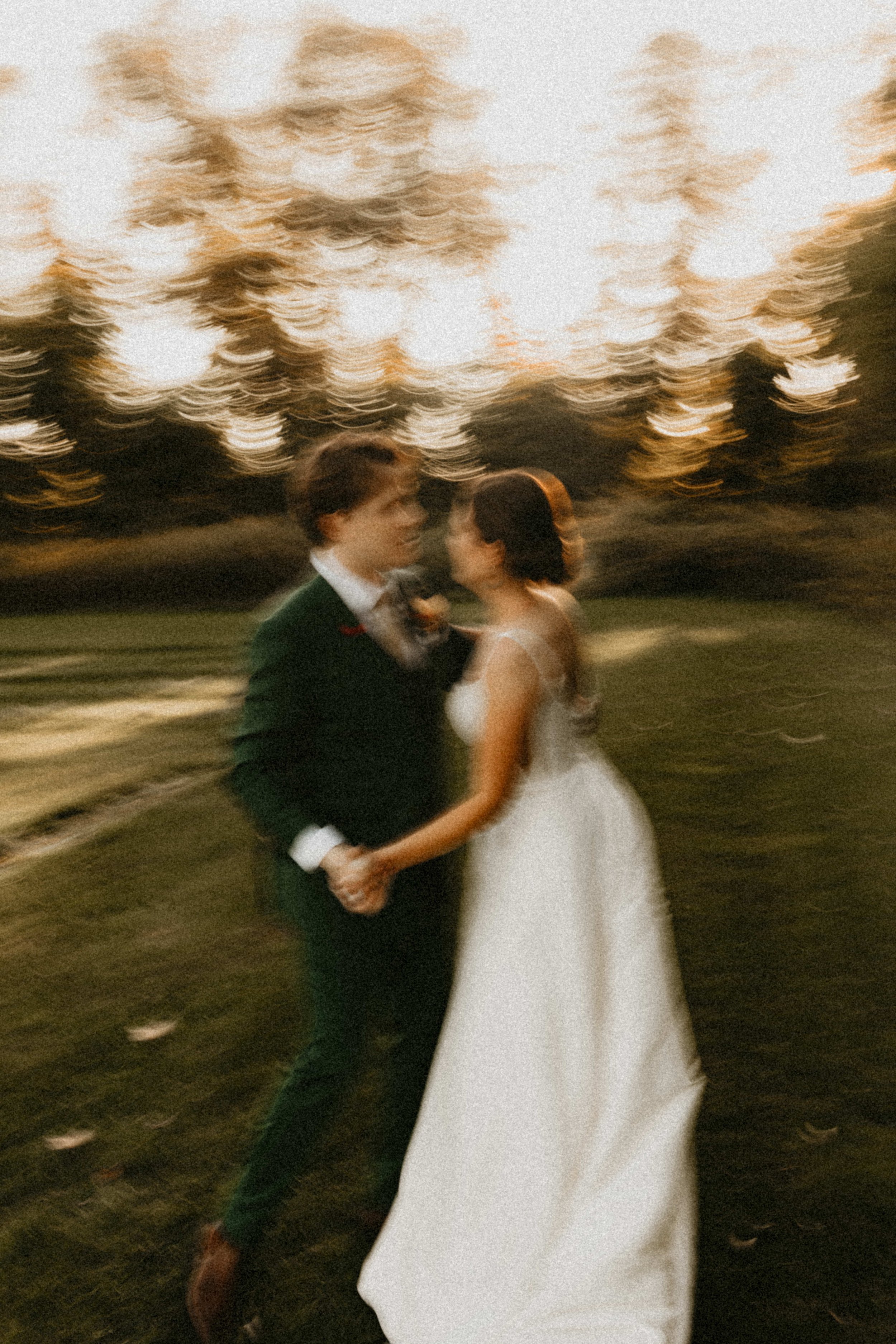 Blurry photo of a young man and woman holding hands, dressed in formal attire, dancing outdoors at sunset with trees in the background.