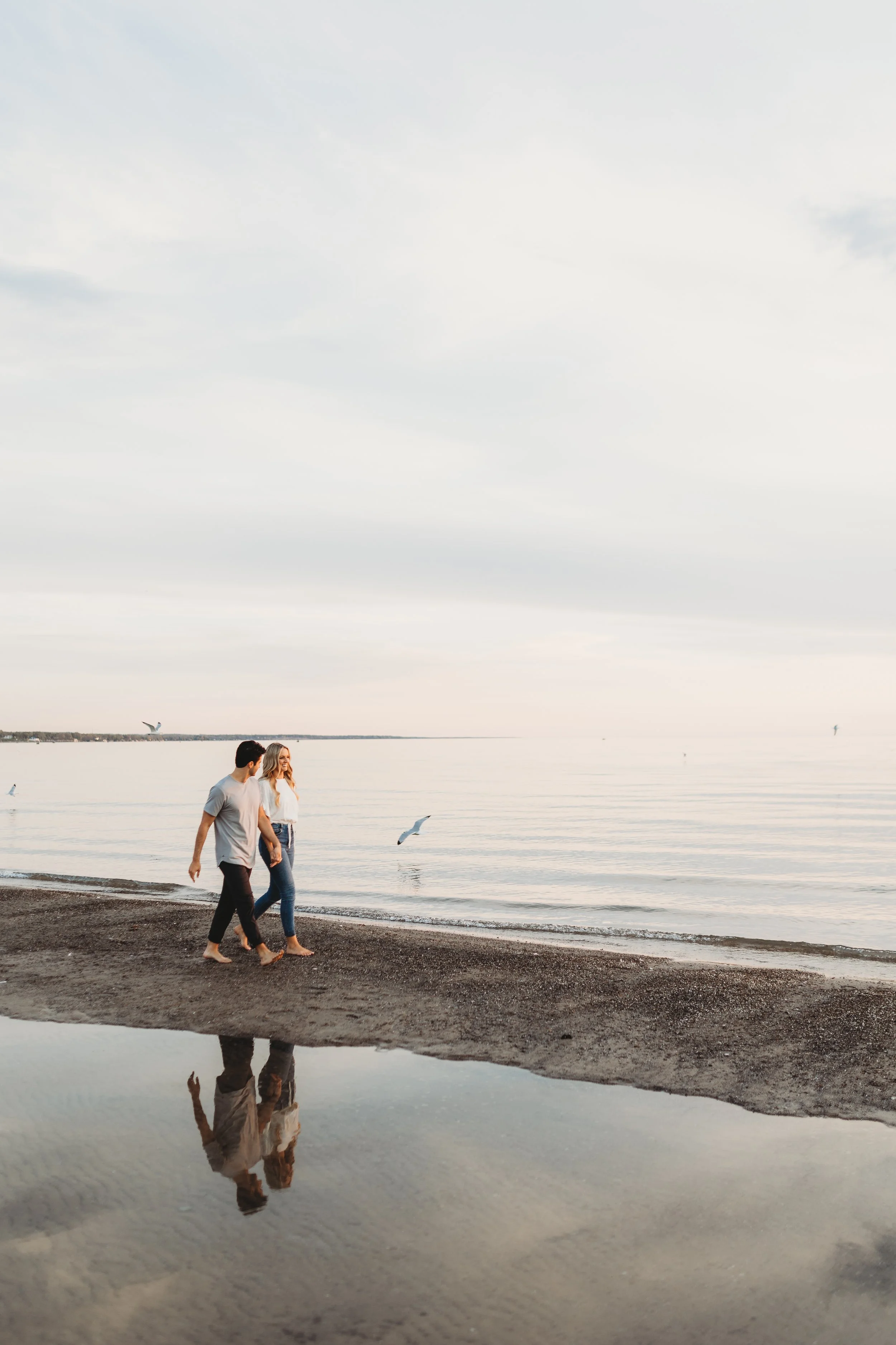 A couple walking along the beach near the water, with seagulls flying nearby, reflected on a small puddle on the sand, under a cloudy sky.