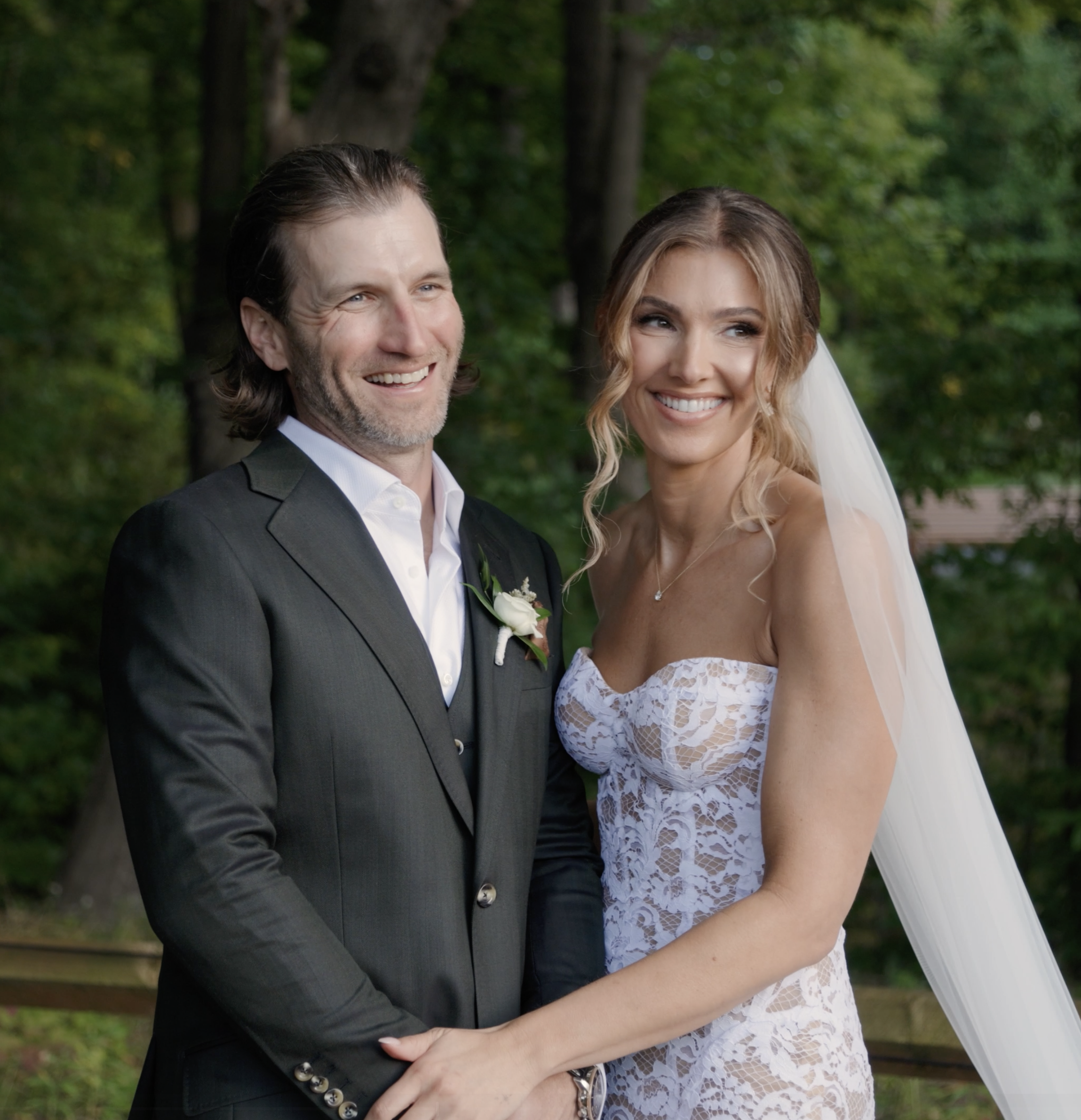 A smiling bride and groom standing close together outdoors, with the bride wearing a white lace wedding dress and veil, and the groom wearing a dark suit, light shirt, and tie, surrounded by green trees.