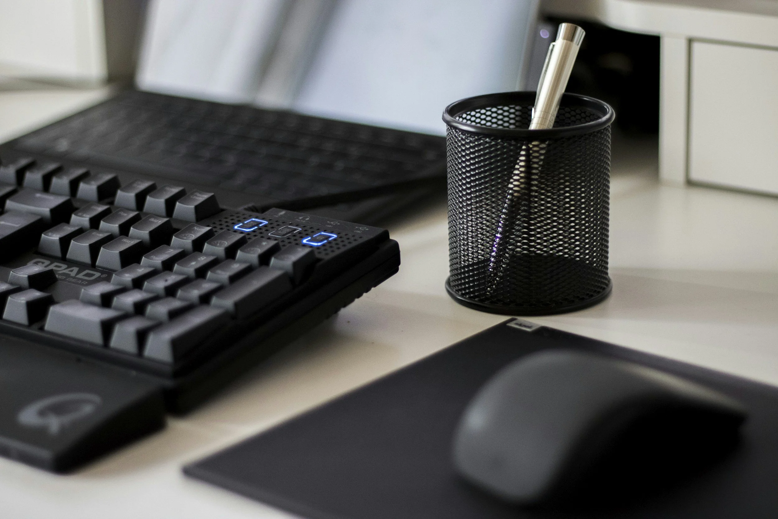 Computer desk setup with a laptop, mechanical keyboard with blue backlights, black mouse on a mouse pad, and a pen in a black mesh pen holder.