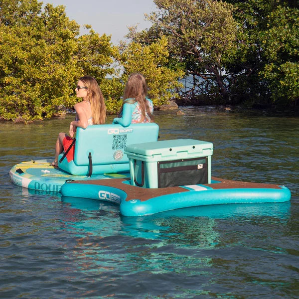 Two girls sitting on a floating inflatable cooler and paddleboard on a lake, with trees in the background.