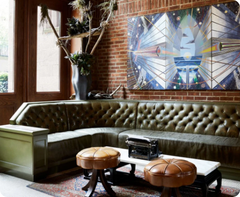 Interior of a cozy living room with a curved olive-green velvet couch, two brown leather footstools, a black vintage radio on a white coffee table, abstract wall art, and a large potted plant near a window.