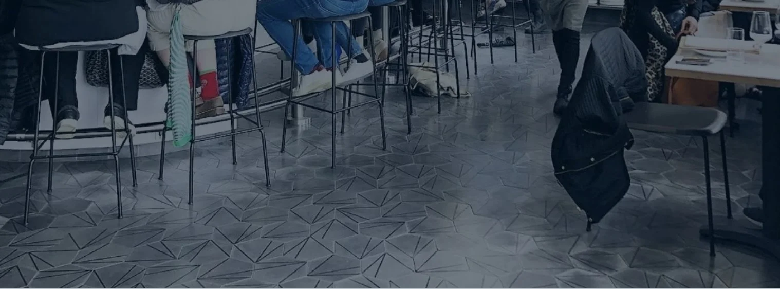 Indoor restaurant or cafe with barstools and tables, some people sitting and some standing, patterned tile floor, and a jacket hanging from a chair.