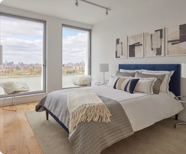 Modern bedroom with a large window, city skyline view, bed with blue headboard, white and striped pillows, beige throw blanket, framed abstract art on the wall, and minimal decor.