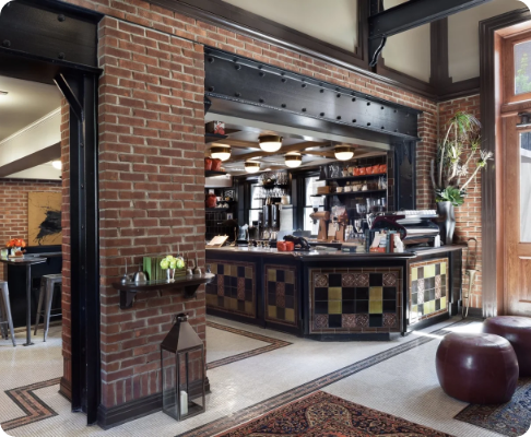 Interior of a cozy cafe or restaurant with a brick wall, a black counter with colorful tile accents, and a large window letting in natural light.